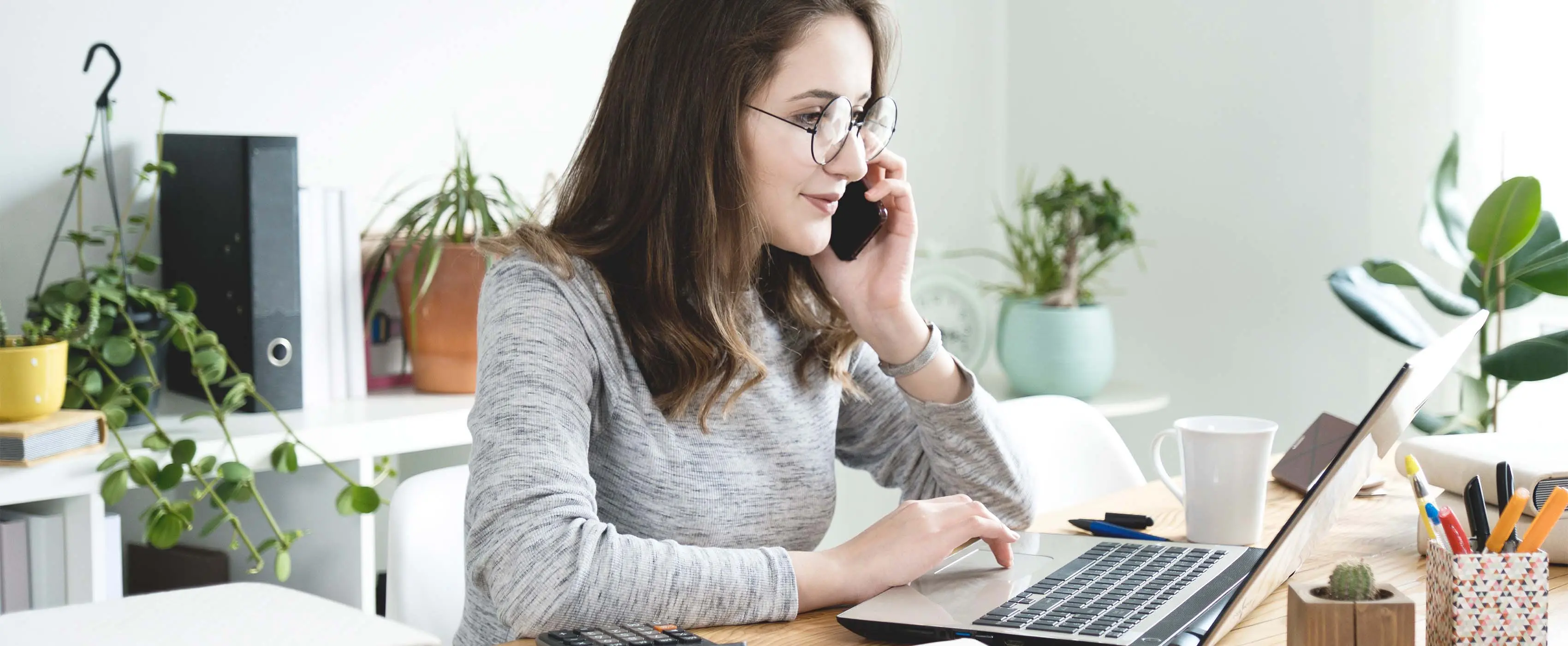 Foto de uma mulher sentada à mesa, falando ao telefone e olhando para a tela de um notebook