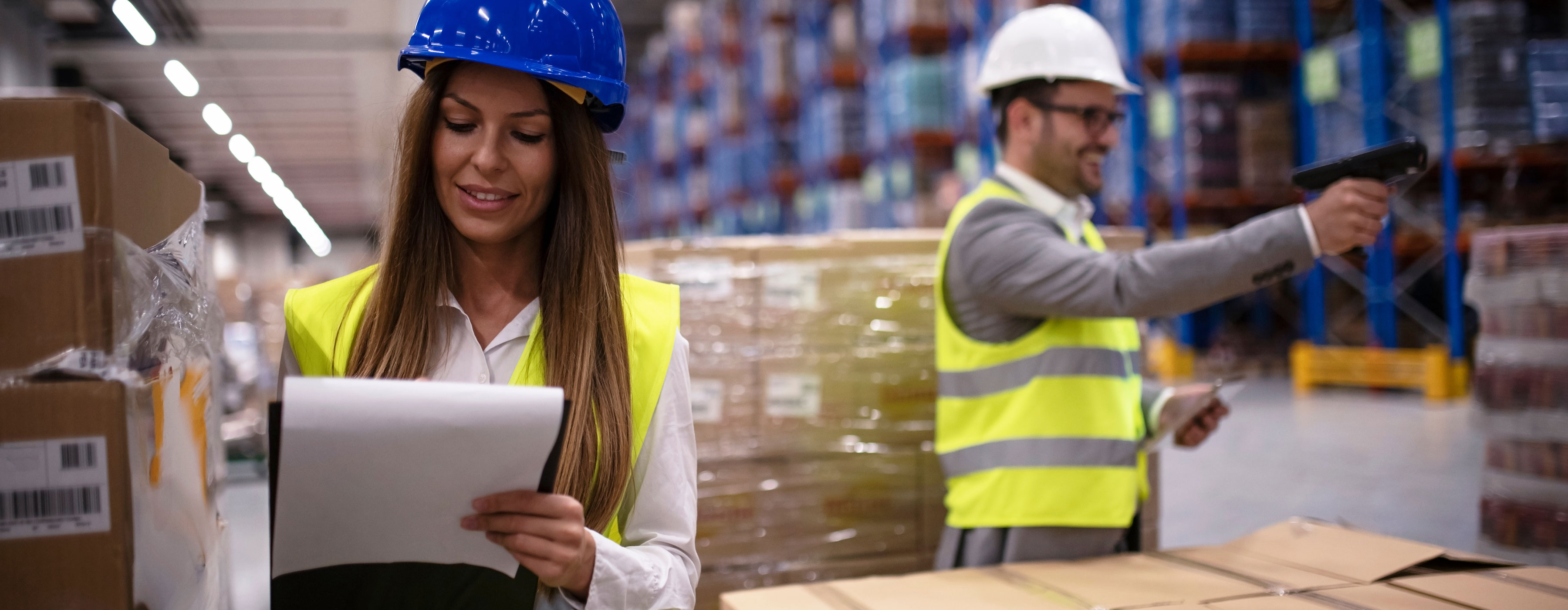 Foto de uma mulher e um homem usando equipamento de segurança em um galpão com prateleiras de cargas. 