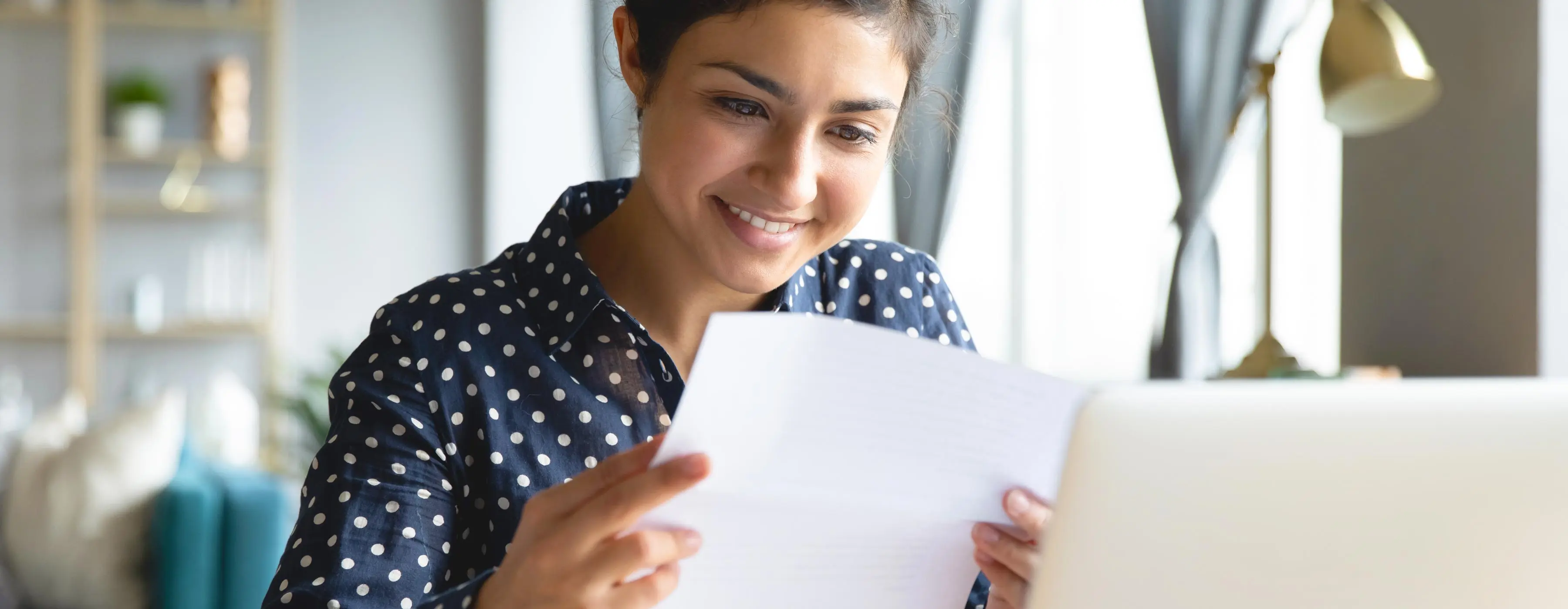 Foto de uma mulher jovem sentada em um ambiente acolhedor sorrindo e verificando um documento em suas mãos.