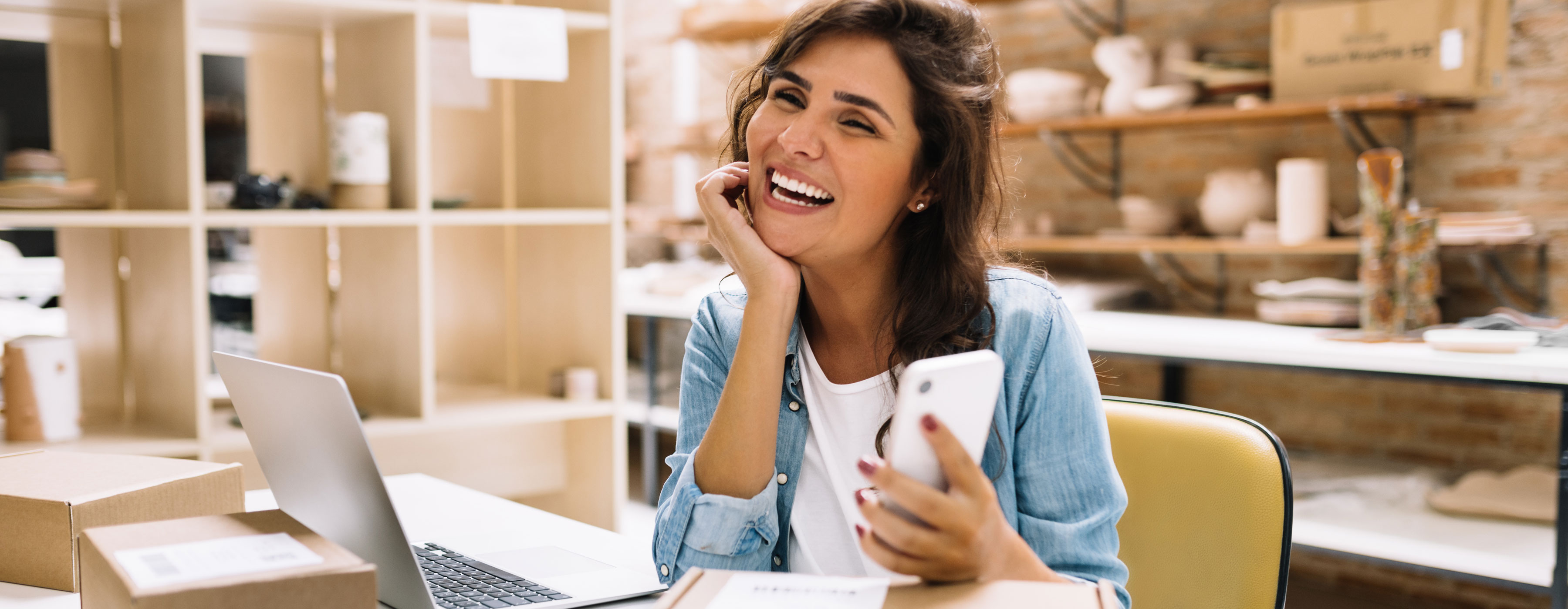 E-commerce: do planejamento à execução - foto de uma mulher sentada à mesa, sorrindo com celular na mão em frente a um laptop.