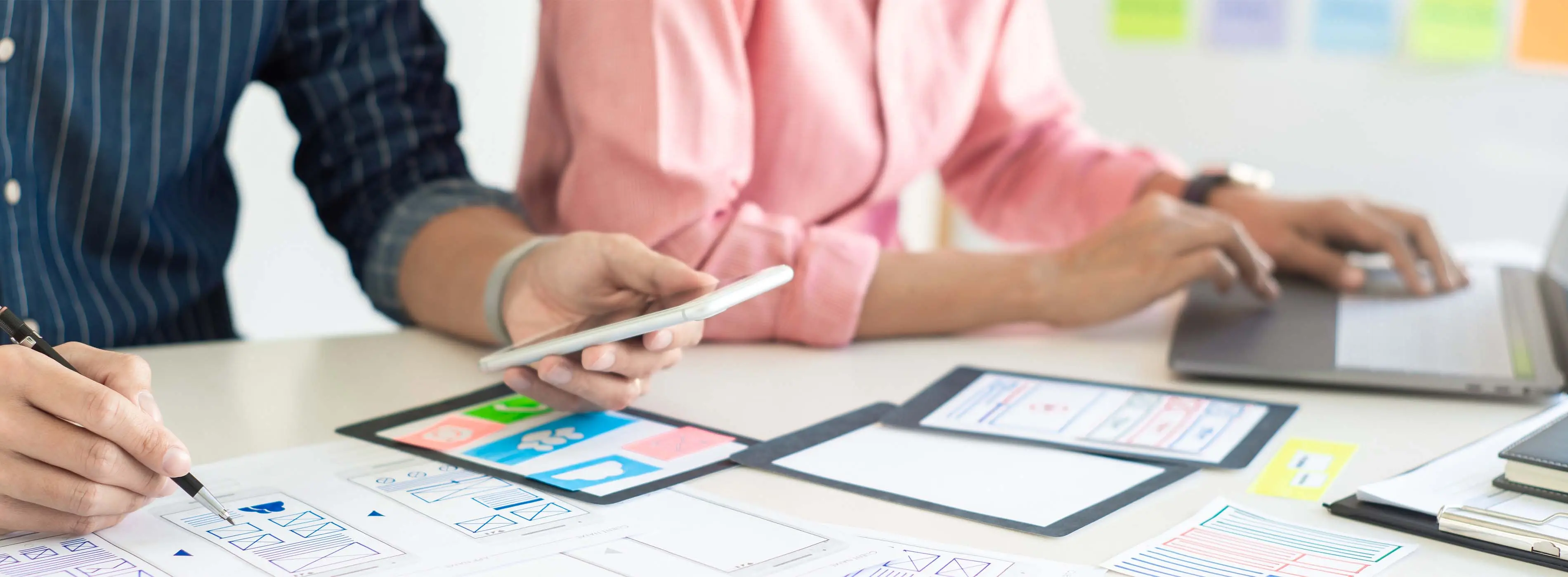 Foto de mãos de duas pessoas, uma pessoa está desenhamdo protótipos de telas de celular e a outra pessoa está utilizando um notebook.