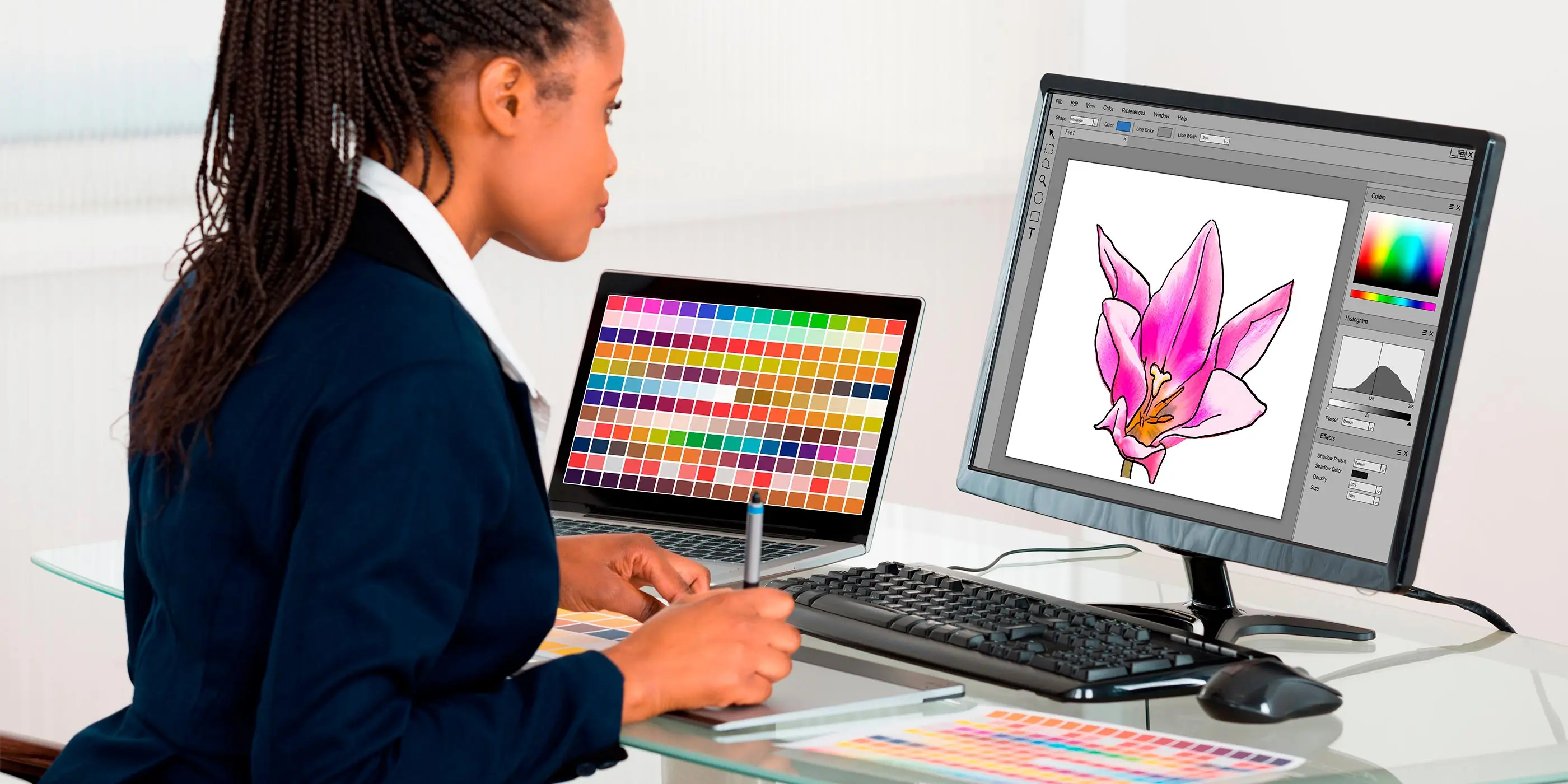 Foto de mulher usando mesa digitalizadora enquanto olha um monitor com desenho de uma flor rosa, e um notebook com paleta de cores na tela ao lado