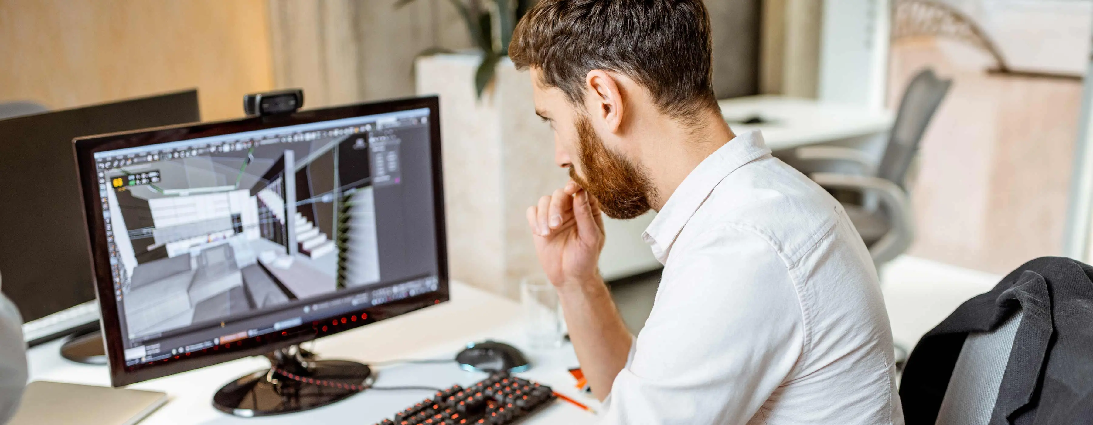 Foto de homem sentado em frente a uma mesa com um computador cuja tela mostra um software de arquitetura.