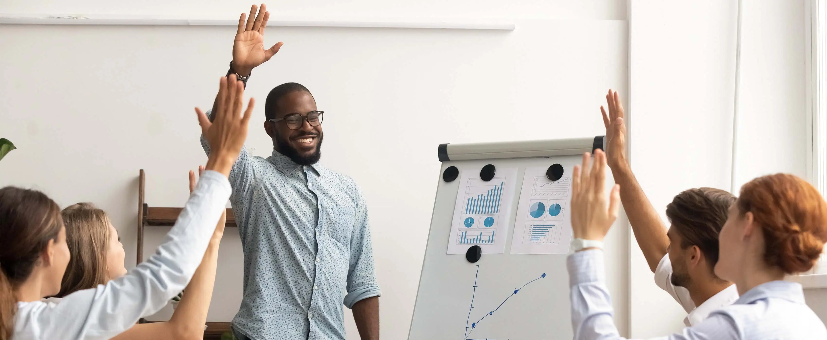 Foto de homem em pé na frente de flip chart com desenhos de gráficos e grupo de pessoas sentadas com mãos levantadas em sala de reunião.