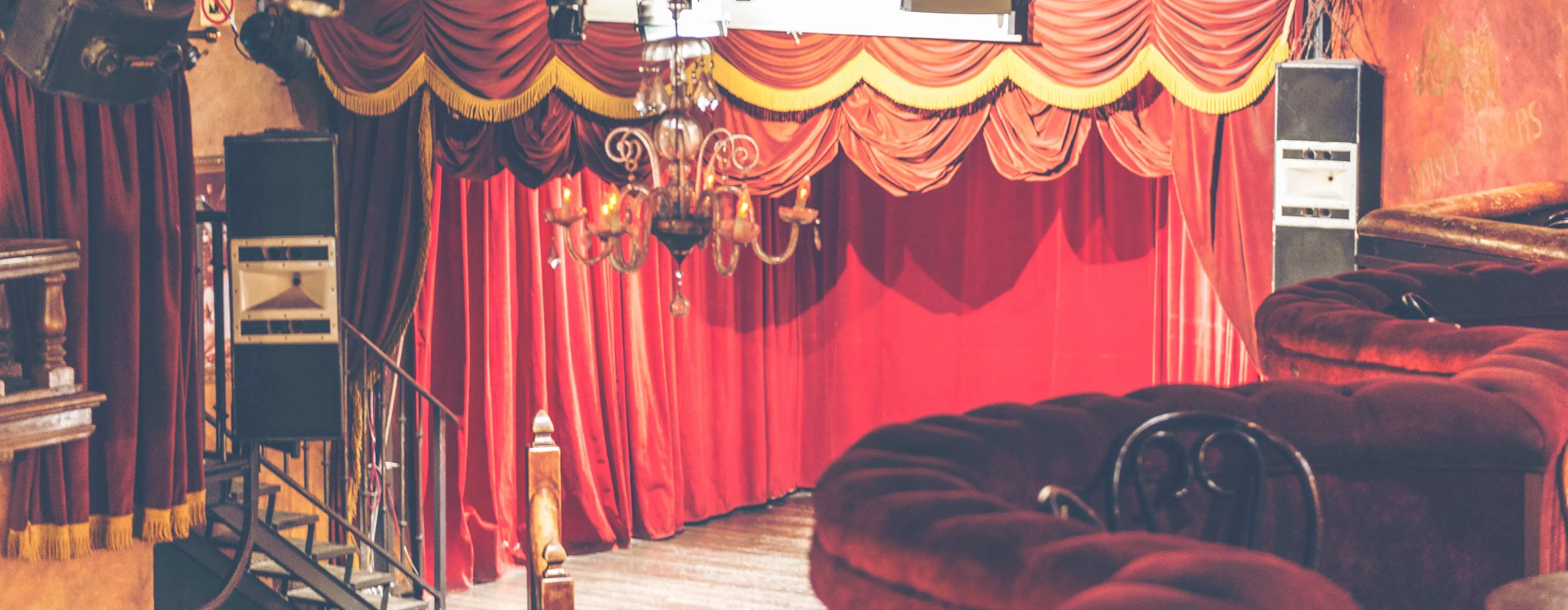 Foto de palco com cortinas vermelhas fechadas, à frente há bancos de veludo vermelho.