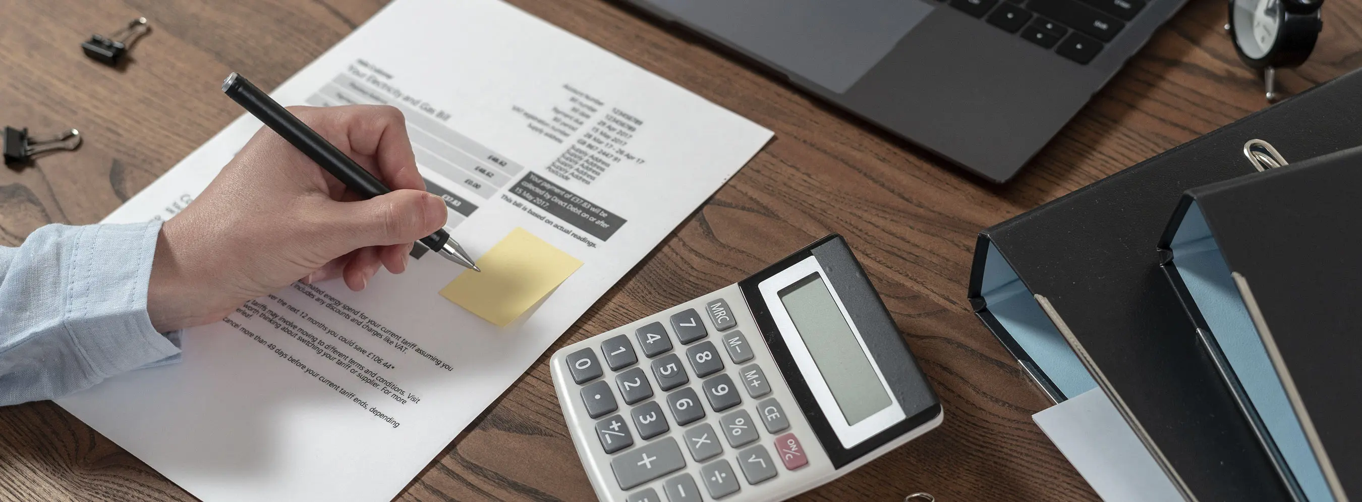 Foto em close de mão de uma pessoa anotando em post it grudado em um papel com tabela em uma mesa com calculadora e fichário e pasta.