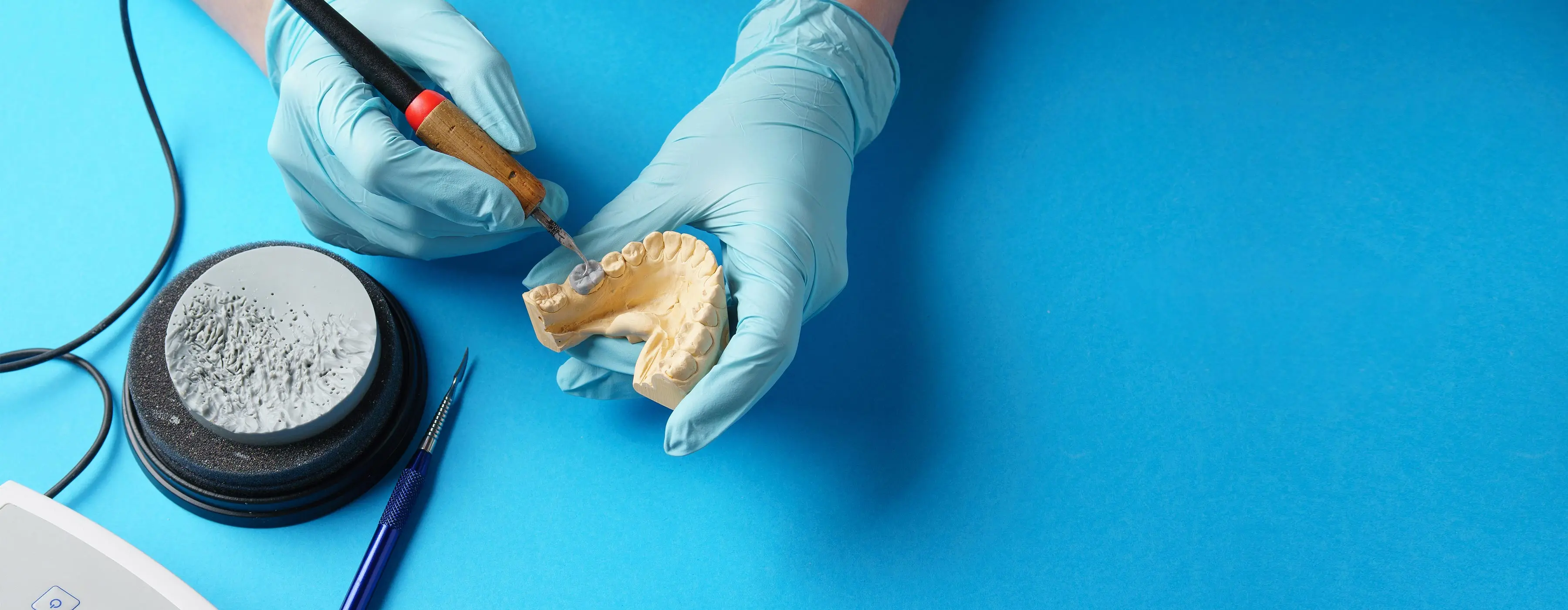 Foto em close de mãos com luvas cirúrgicas manipulando uma prótese dentária utilizando ferramentas de odontologia.