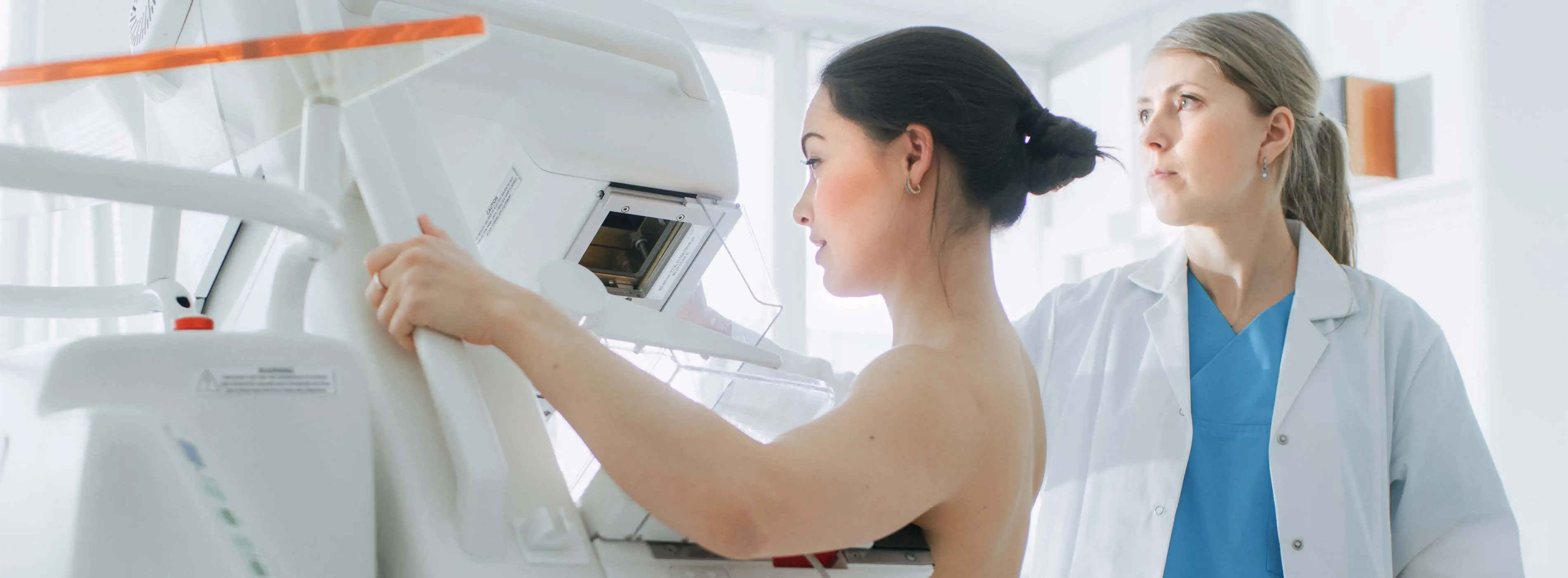 Foto de uma mulher fazendo mamografia, com outra mulher, com roupas de médica, ao lado e manipulando o equipamento do exame.