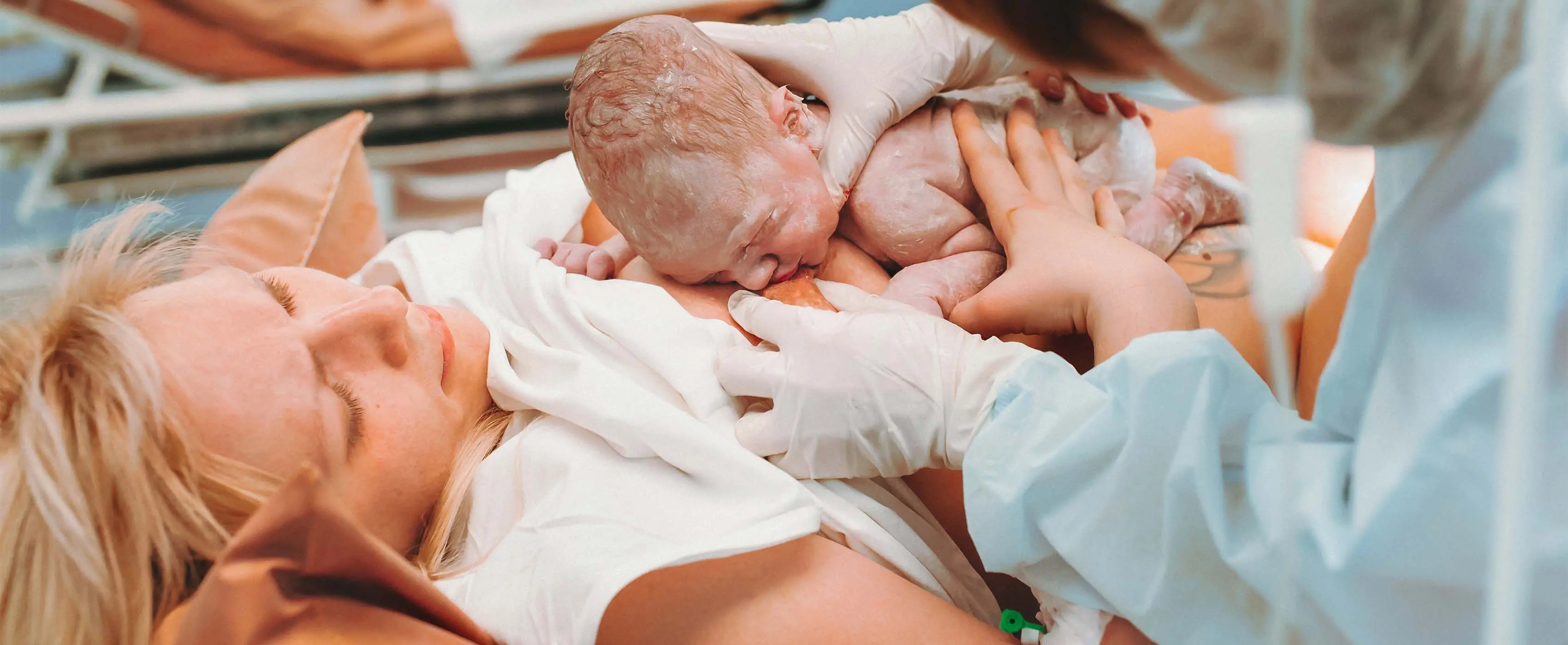 Foto de uma enfermeira auxiliando uma mulher deitada com o bebe recém-nascido em seu seio.