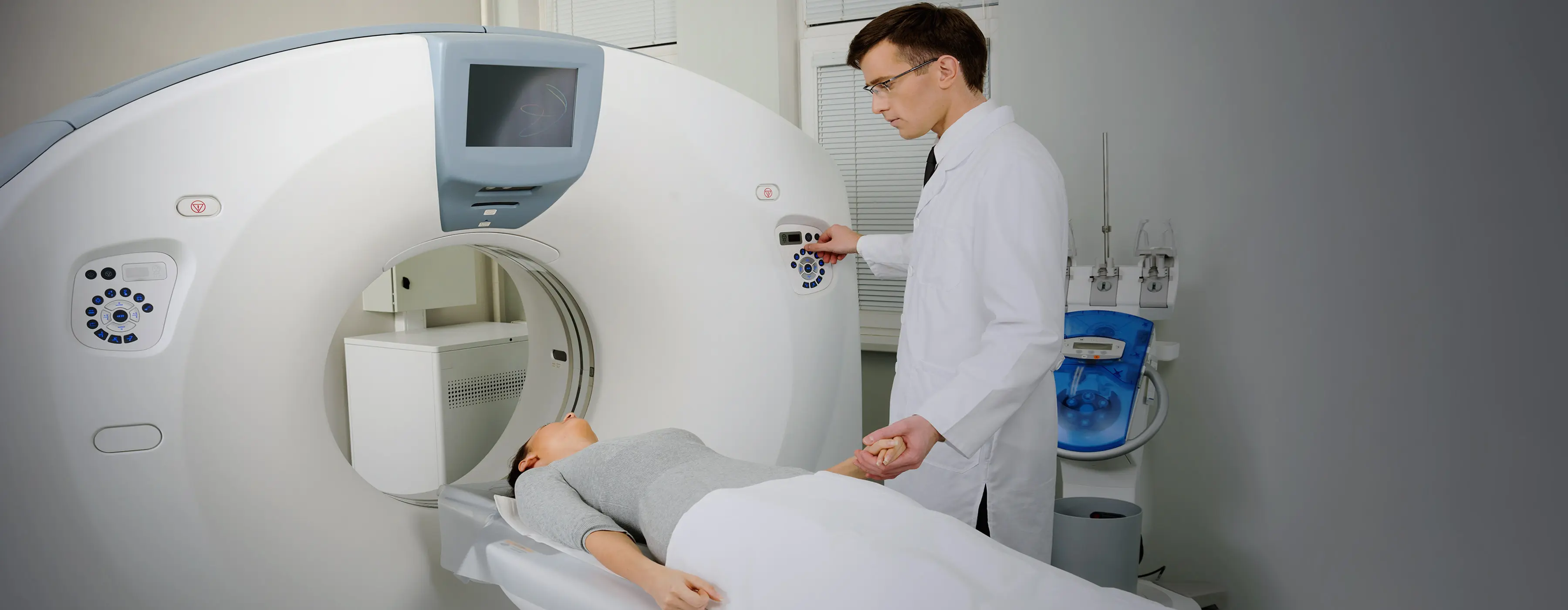 Foto de mulher deitada em uma máquina de tomografia que está sendo manipulada por um homem vestido com jaleco branco.