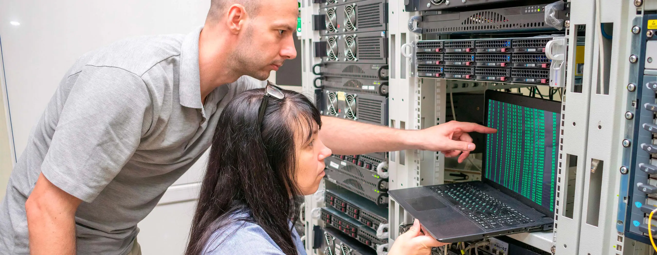 Foto de um homem e uma mulher em uma sala de servidor de informática verificando sistemas em um notebook.