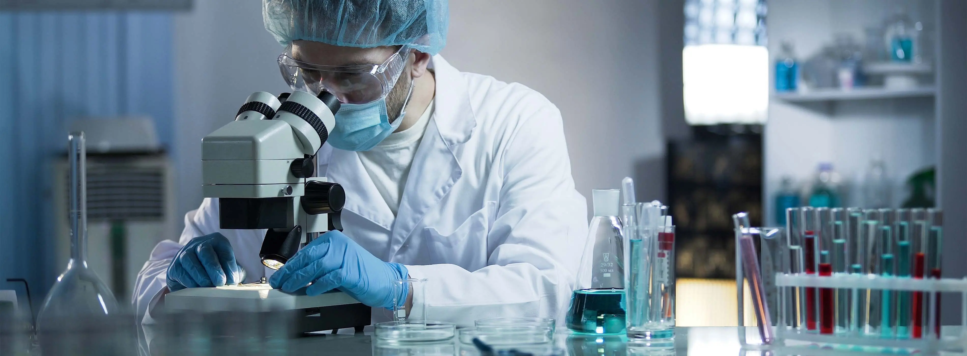 Foto de uma pessoa com jaleco, luvas cirúrgicas, máscara, touca e óculos de proteção manipulando um microscópio em um cenário de laboratório clínico.