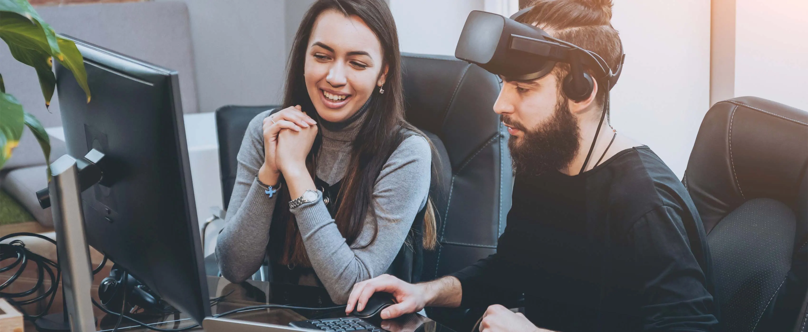 Foto de um homem usando óculos de realidade virtual mexendo em um computador e conversando com uma mulher que observa a tela do computador.