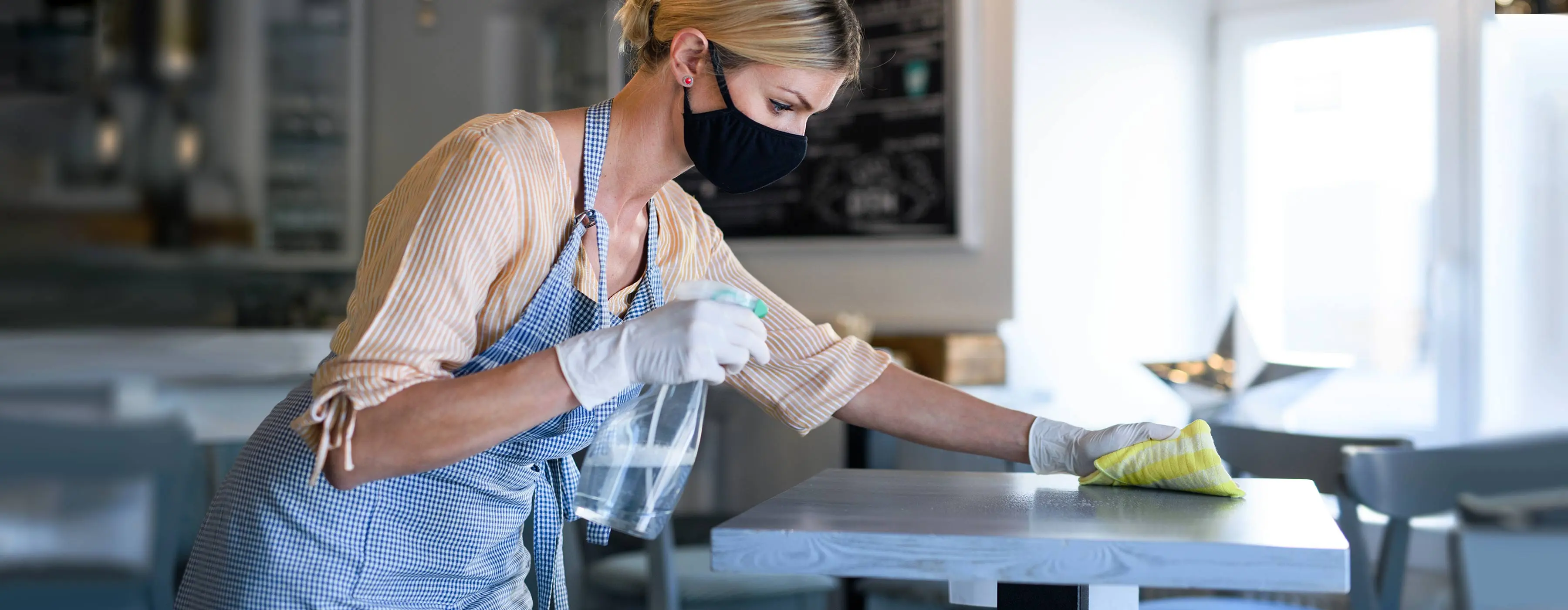 Foto de mulher com avental e máscara limpando mesa com pano e spray em restaurante.