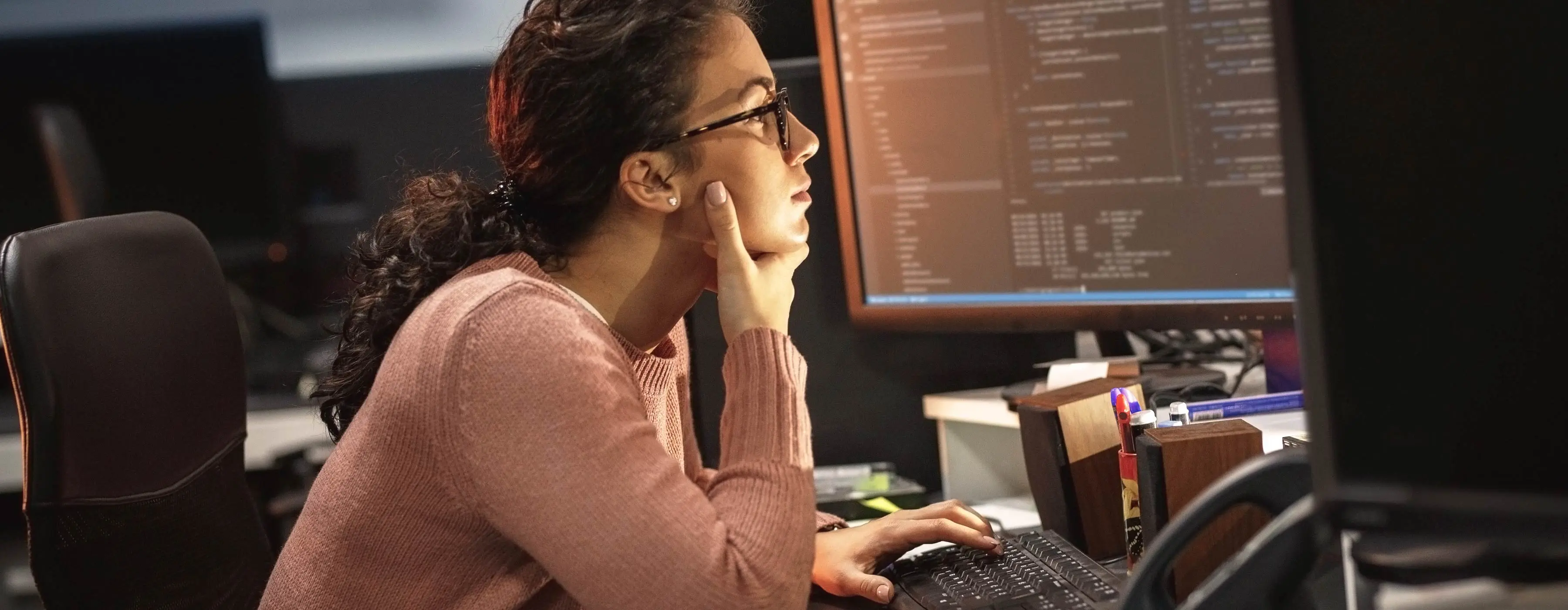 Foto de jovem sentada em escritório, observando códigos em tela de computador.