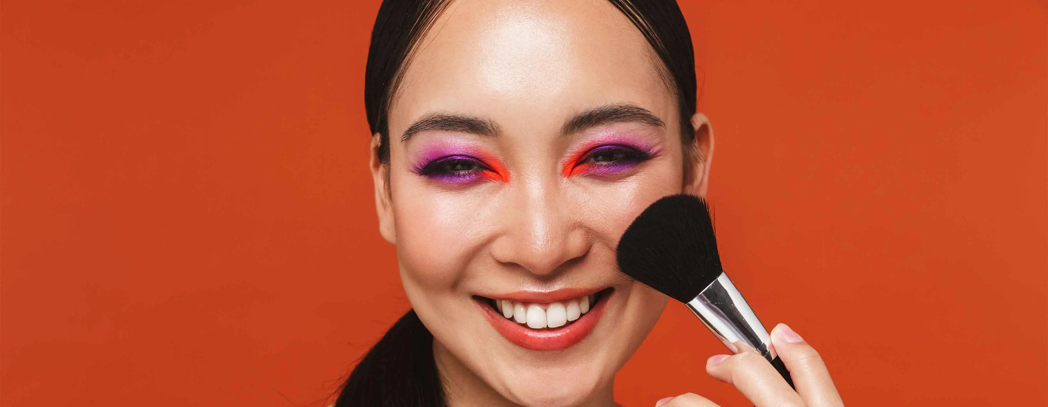 Foto de uma mulher com sombra roxa e vermelha nos olhos, segurando um pincel de blush.