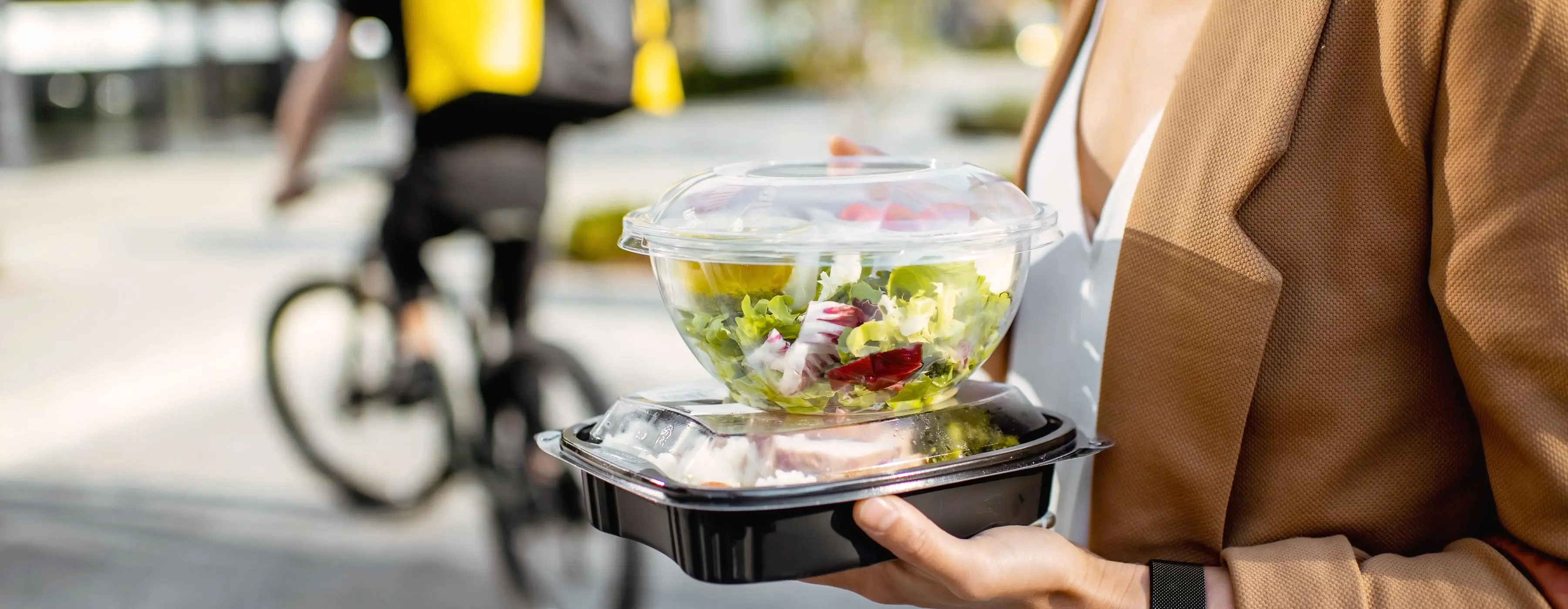 Foto de mulher segurando duas marmitas: uma salada e outra refeição saudável. Ela está na rua e ao fundo ciclista entregador de comida.