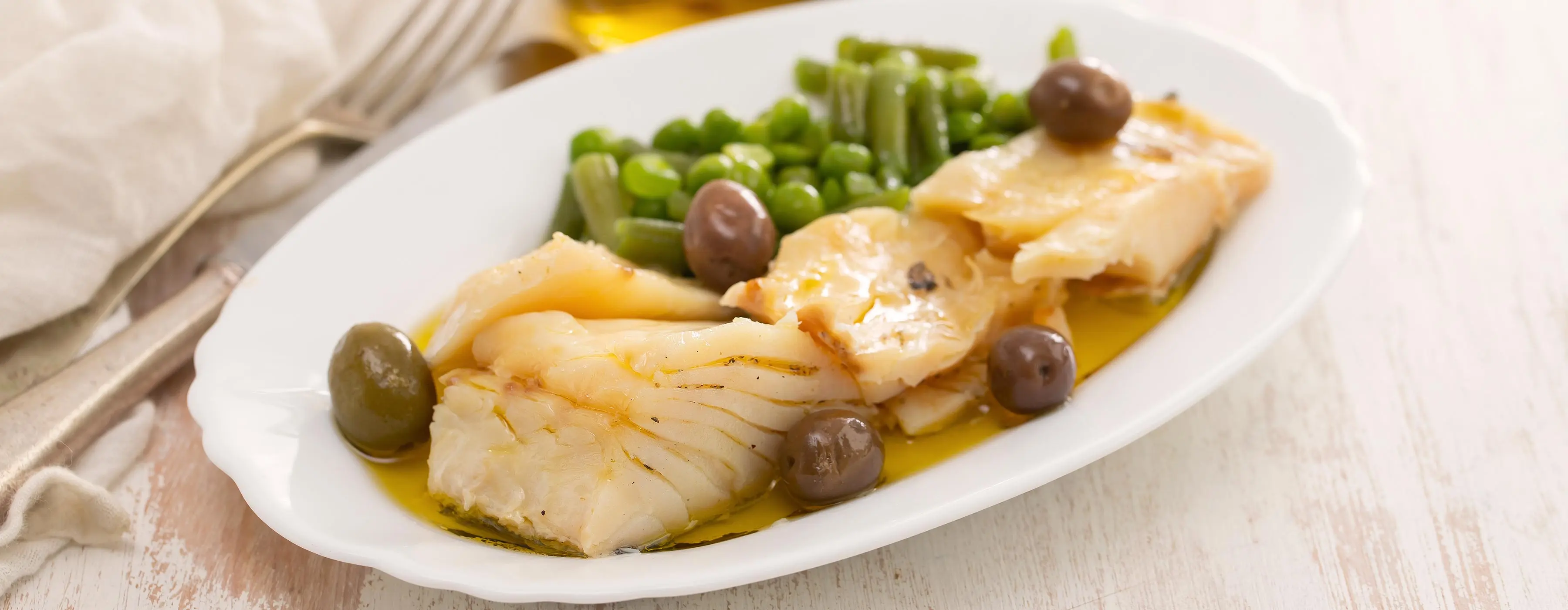 Foto de bacalhau cozido e servido em prato com vagem e azeitonas em mesa com talheres e guardanapo.