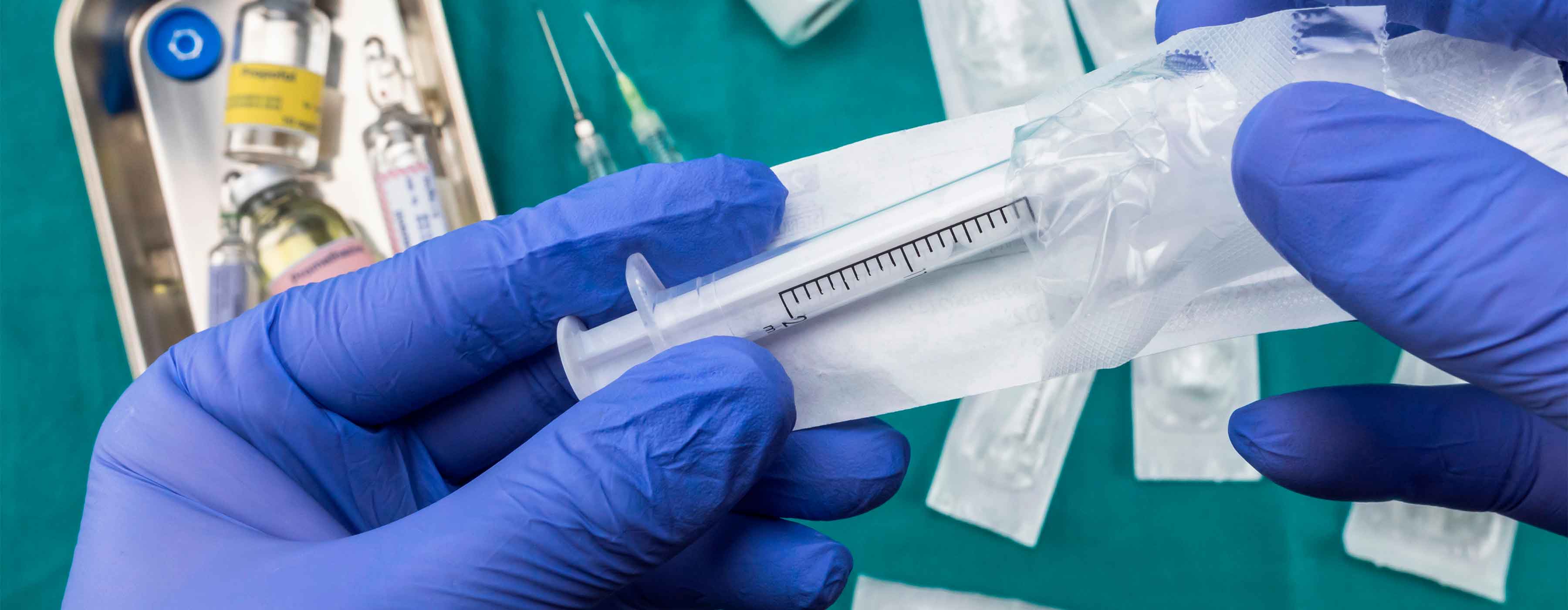 Mãos com luvas azuis abrindo a embalagem de uma seringa descartável, com frascos de medicamentos, agulhas e outros materiais hospitalares ao fundo, sobre uma superfície verde.
