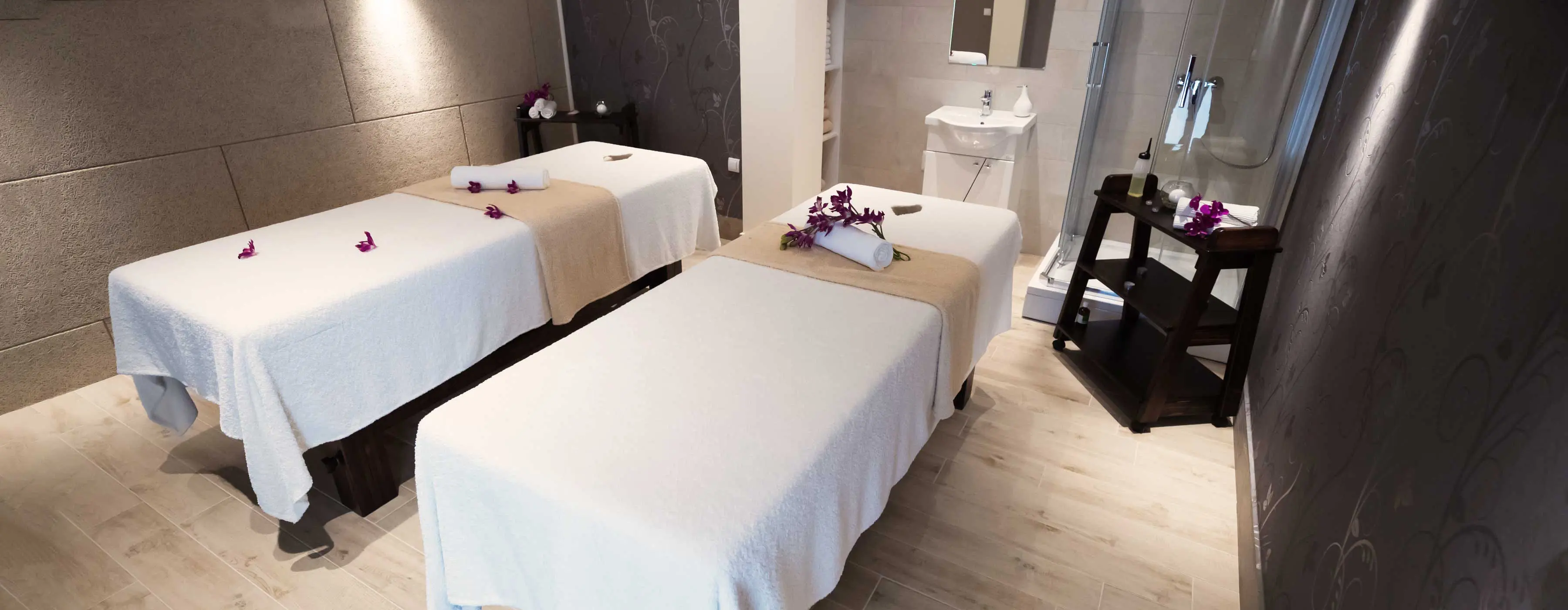 Foto de duas macas de massagem enfeitadas com toalhas e flores em sala de massagem com pia e ducha. 
