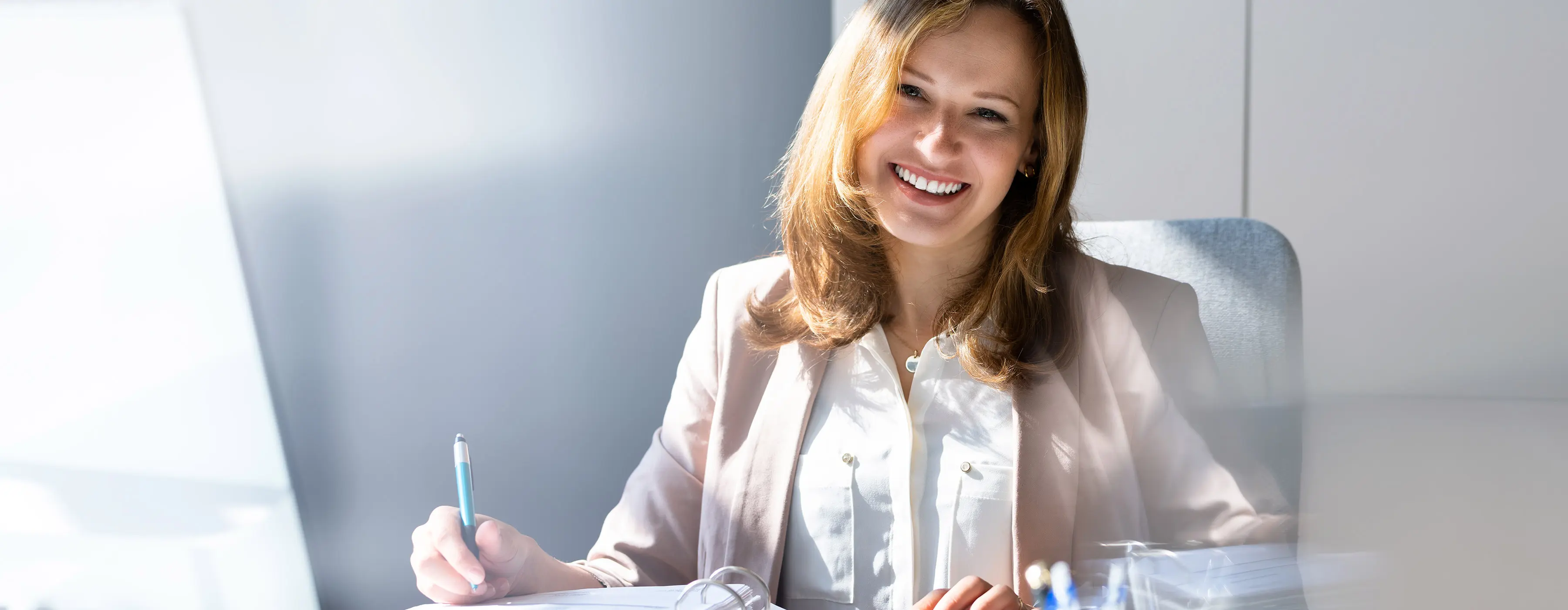 Foto de mulher sorrindo sentada em escritório calculando em máquina de calculadora e a notando em fichário.