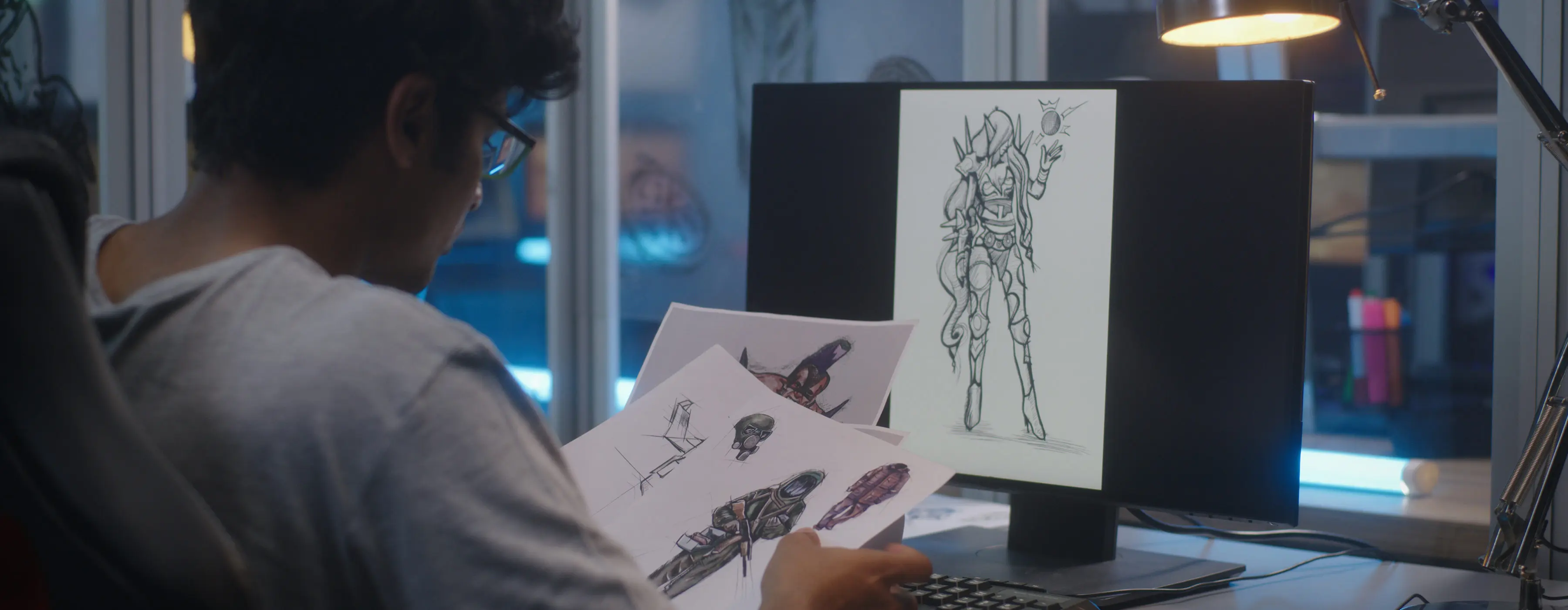 Foto de um homem sentado em frente ao computador e segurando folhas de papeis com ilustrações desenhadas.