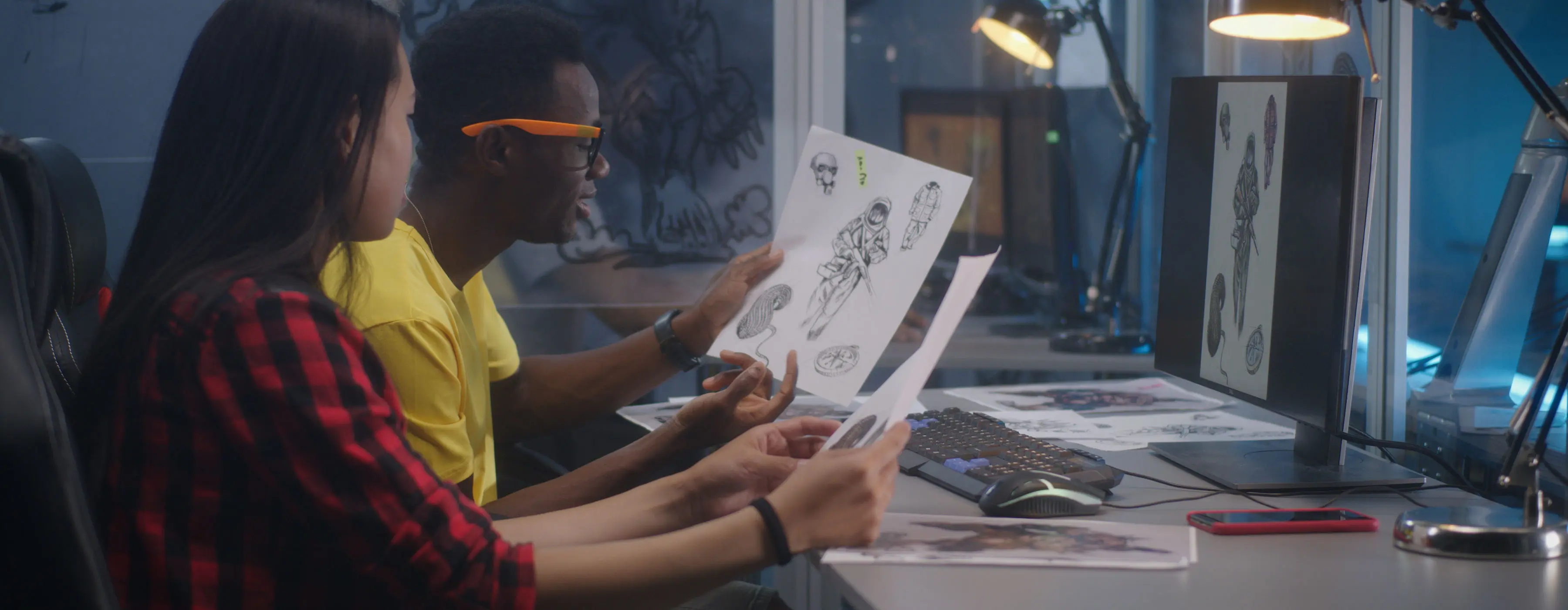 Foto de uma mulher e um homem sentados em frente ao computador e olhando ilustrações feitas no papel.
