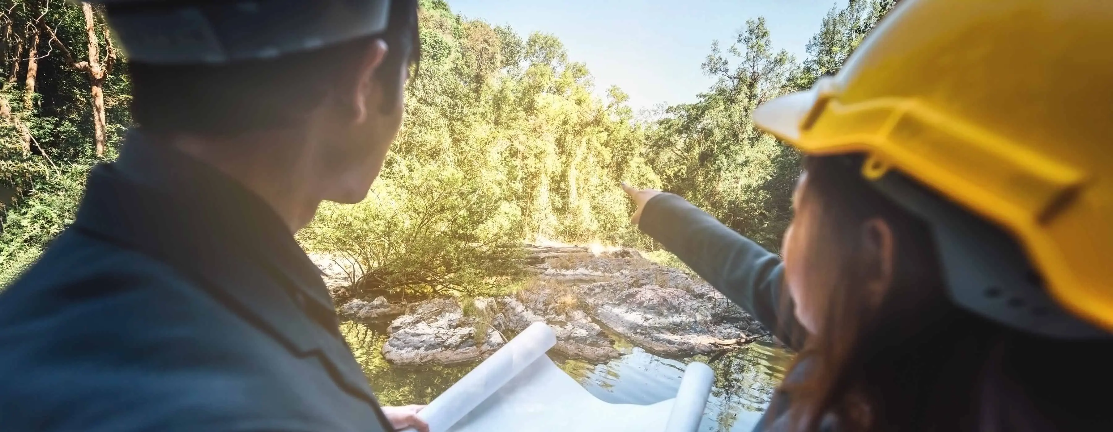 Foto de duas pessoas usando capacete de proteção apontando para o horizonte em uma floresta em cima de uma pedra.
