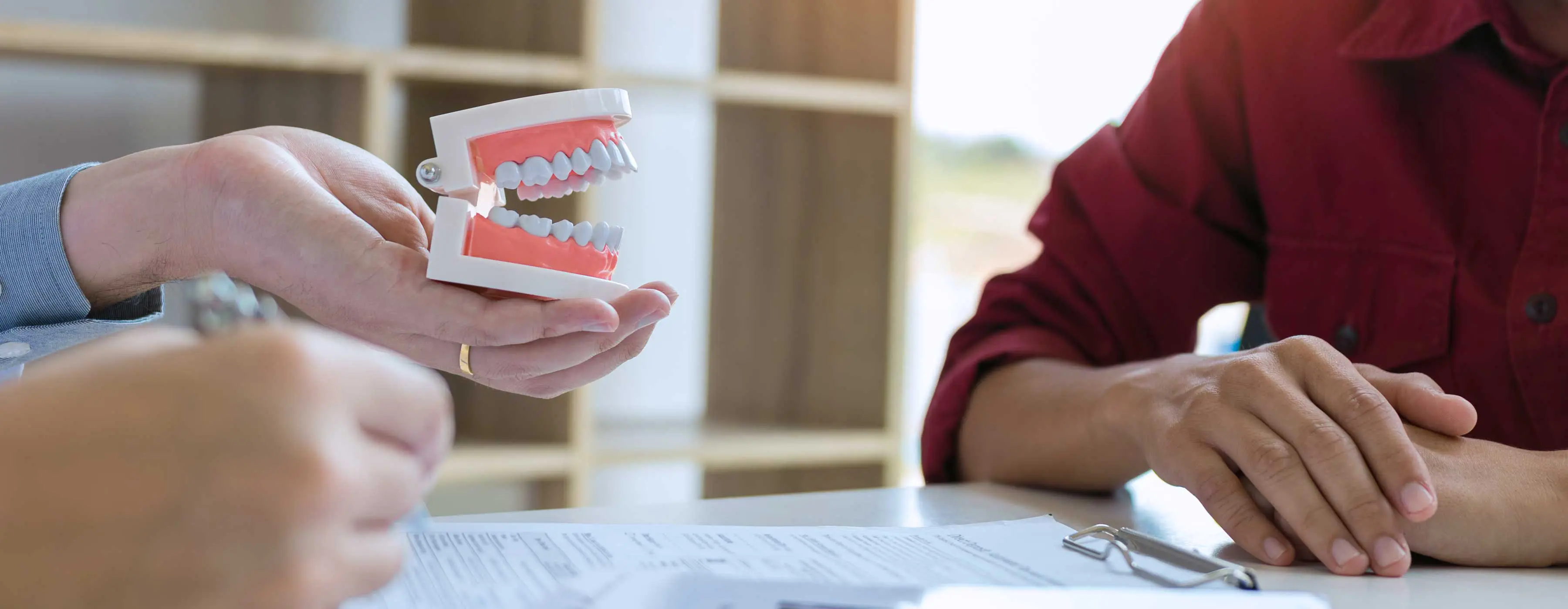 Foto de uma pessoa preenchendo formulário e na outra mão segura arcada dentária e mostra para outra pessoa também sentada em consultório.