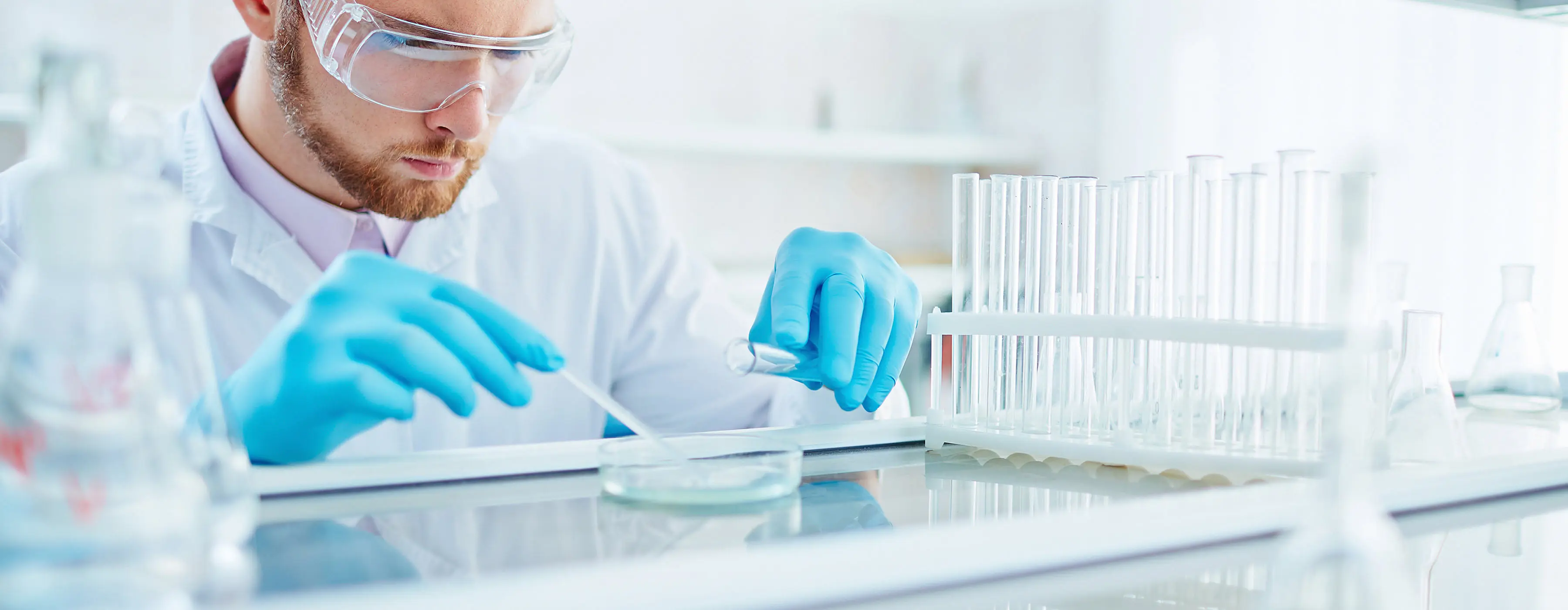 Foto de homem observando amostra em laboratório, usando luvas e óculos de proteção.