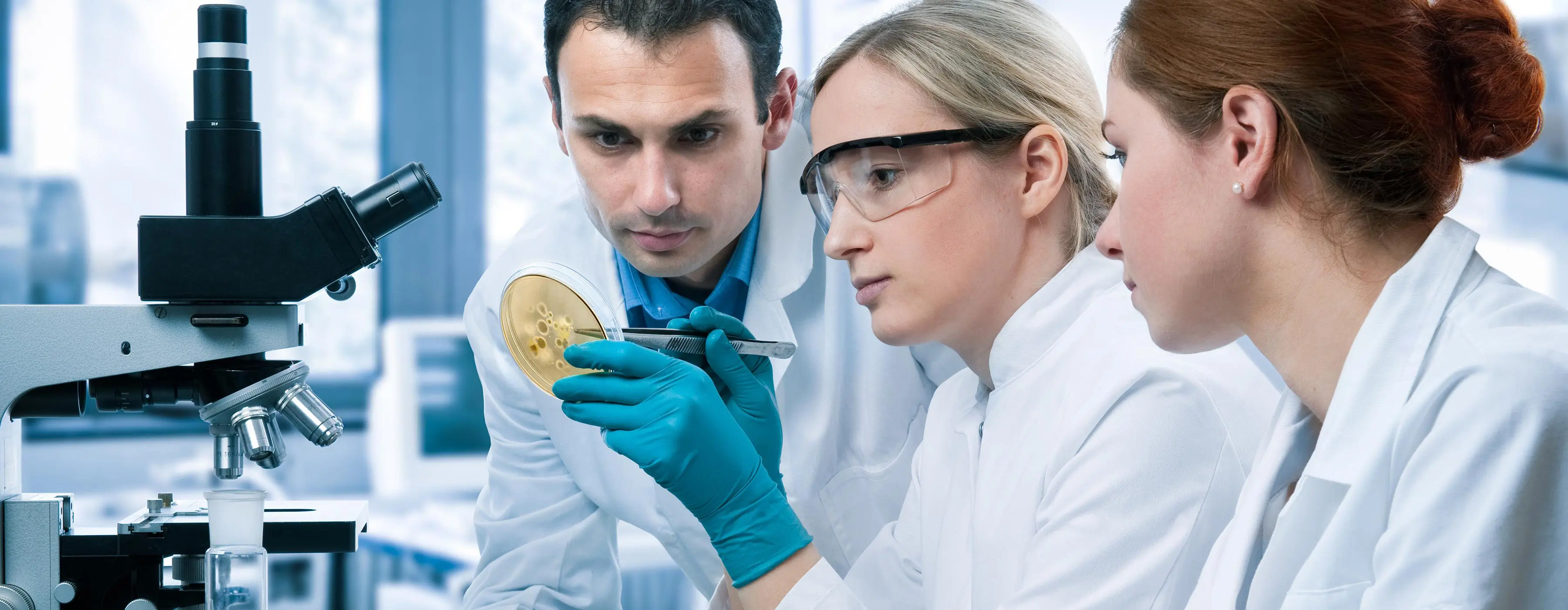 Foto de três pessoas observando pote com amostra de substâncias e uma delas anota em prancheta em laboratório.