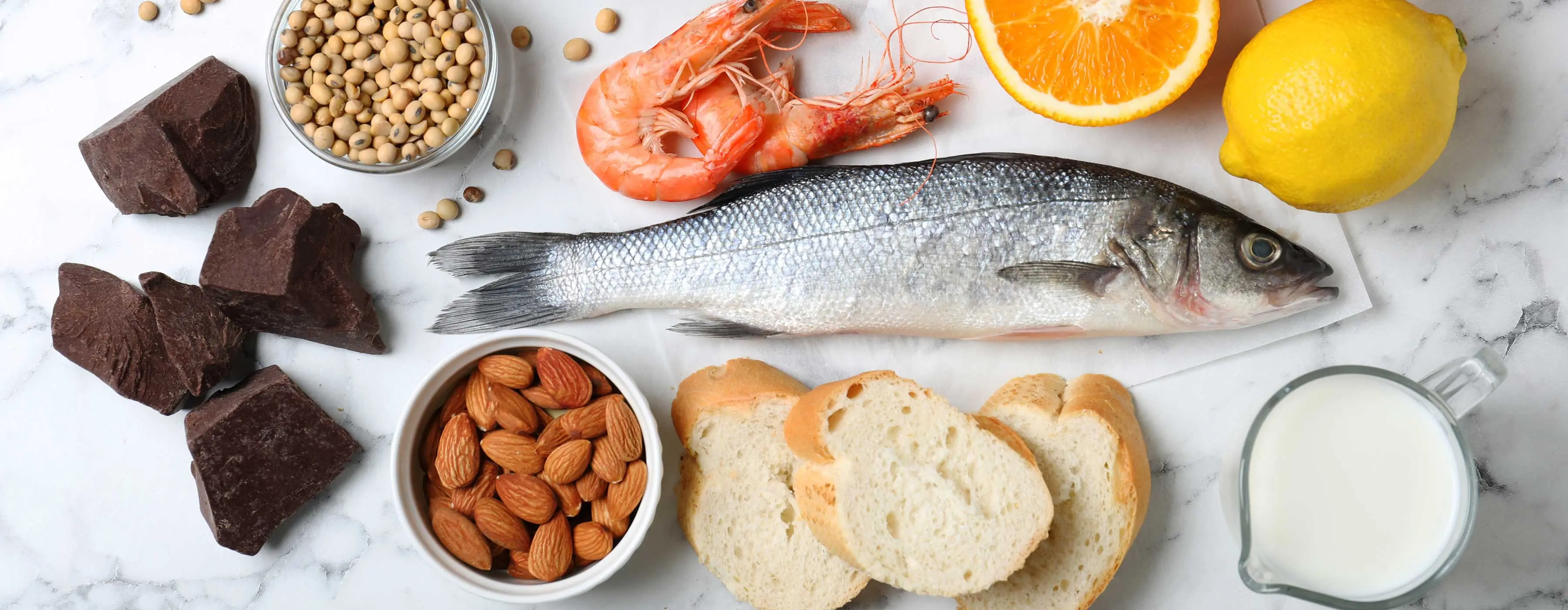 Foto de mesa de mármore em tons de branco e cinza com peixe, camarão, fatias de pão, amêndoas, leite, chocolate, grãos, laranja e limão siciliano.