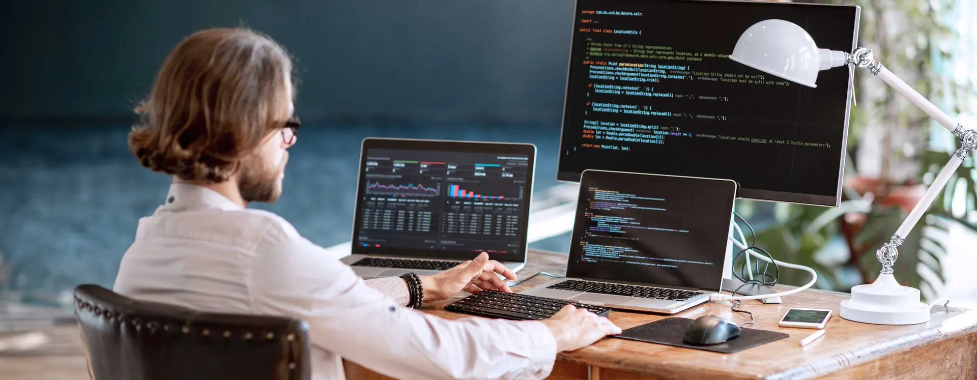 Foto de uma mesa rústica de madeira no escritório com um profissional sentado em frente a três monitores contendo códigos de programação