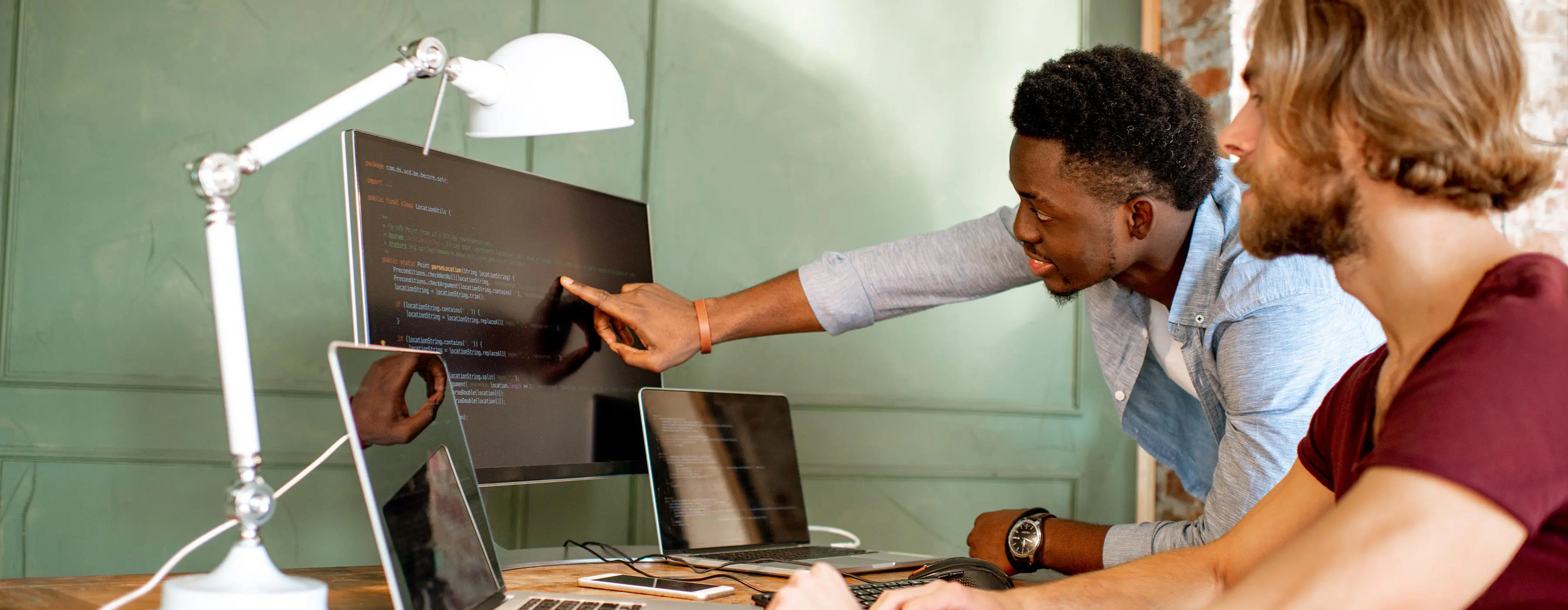 Foto de dois homens olhando tela de computador em software de programação de sites, junto com dois notebooks.
