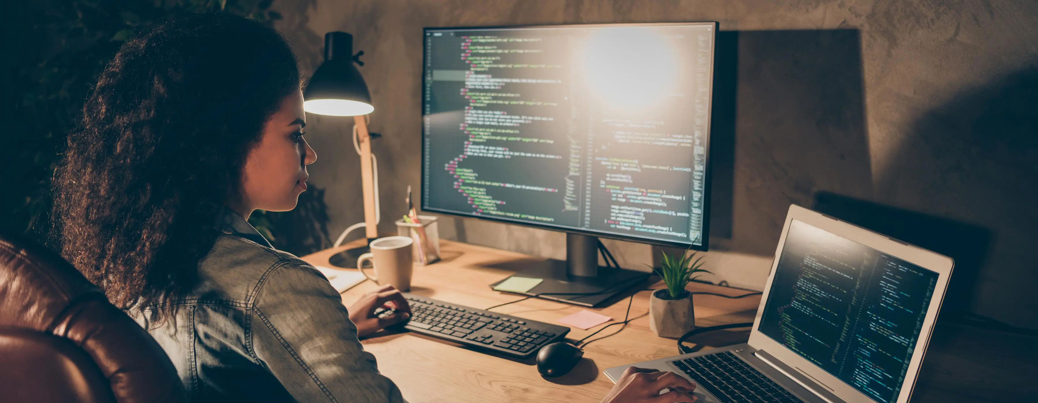 Foto de uma mulher desenvolvedora de software usando computador e notebook testando código em sistema operacional.