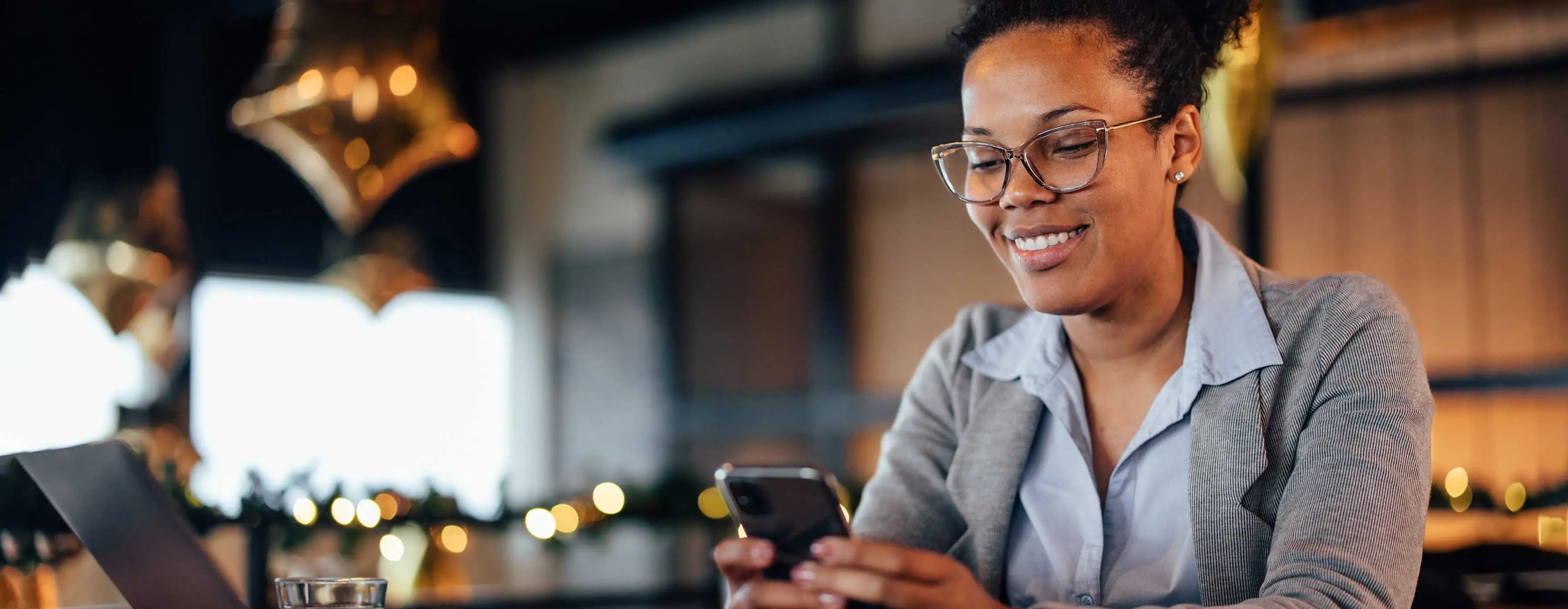 Foto de mulher com camisa, terno e óculos sorrindo ao olhar para celular entre as mãos.