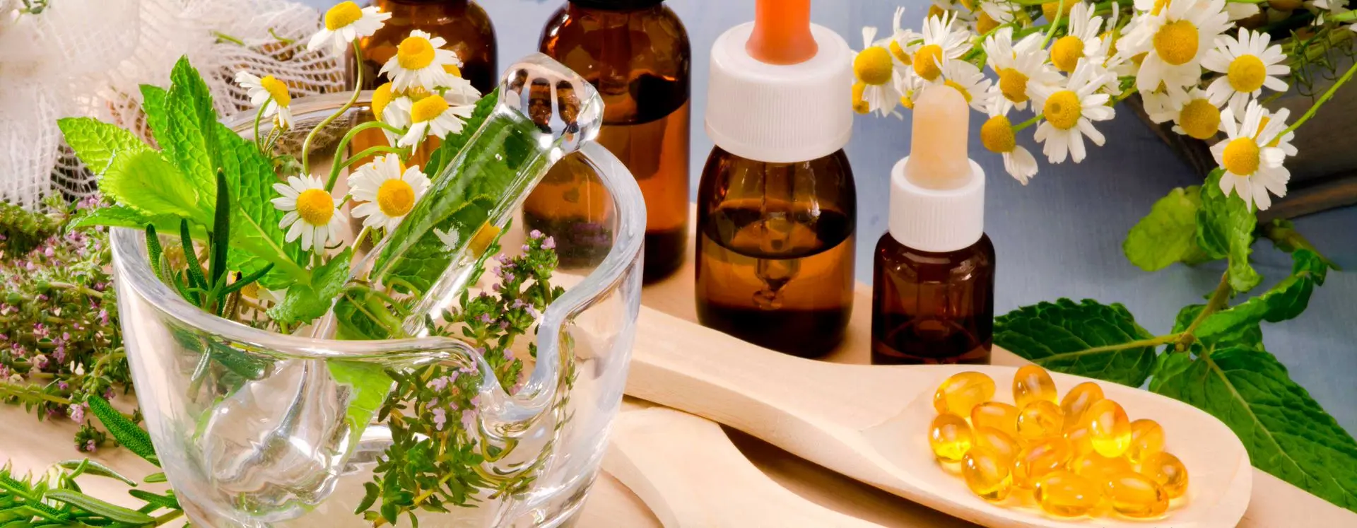 Mesa com flores e vidros de medicação em diversos tamanhos, todos com conta-gotas de borracha na tampa. Também há uma jarra grande de cristal com ervas e flores dentro.