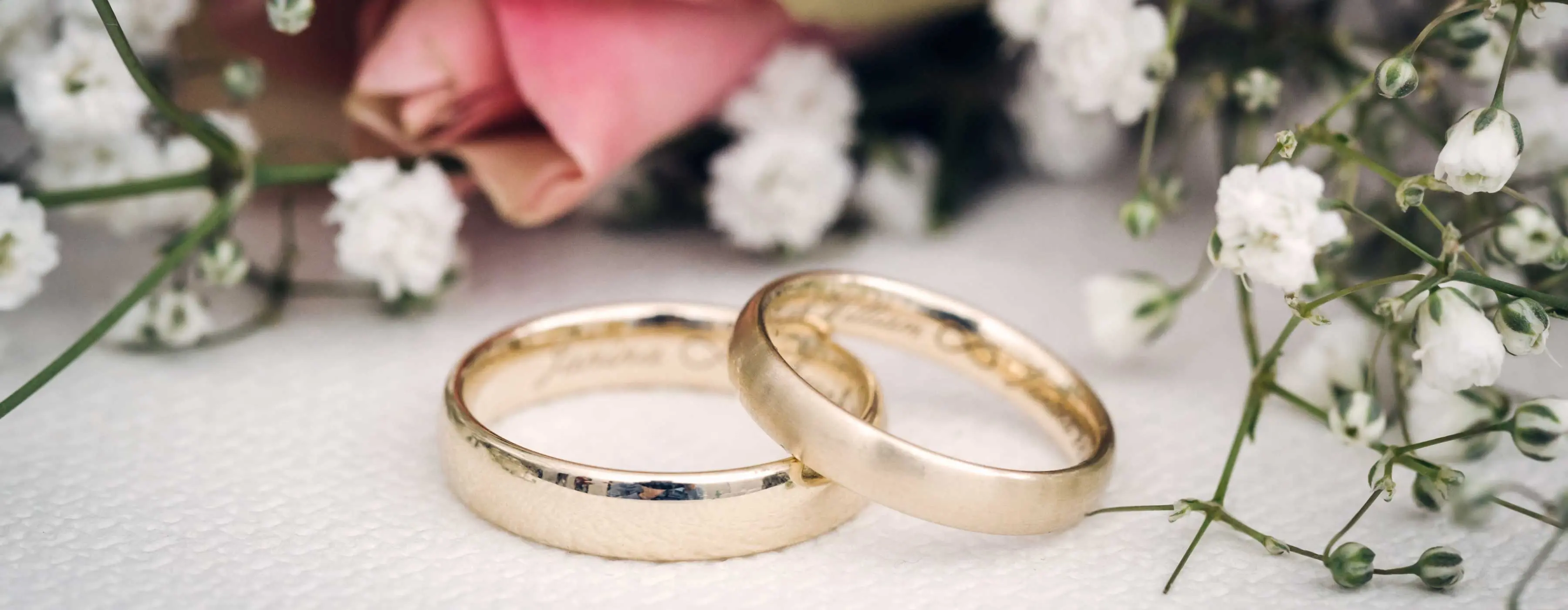 Foto em close para aliança de matrimônio com nomes gravados e flores em cima de mesa.
