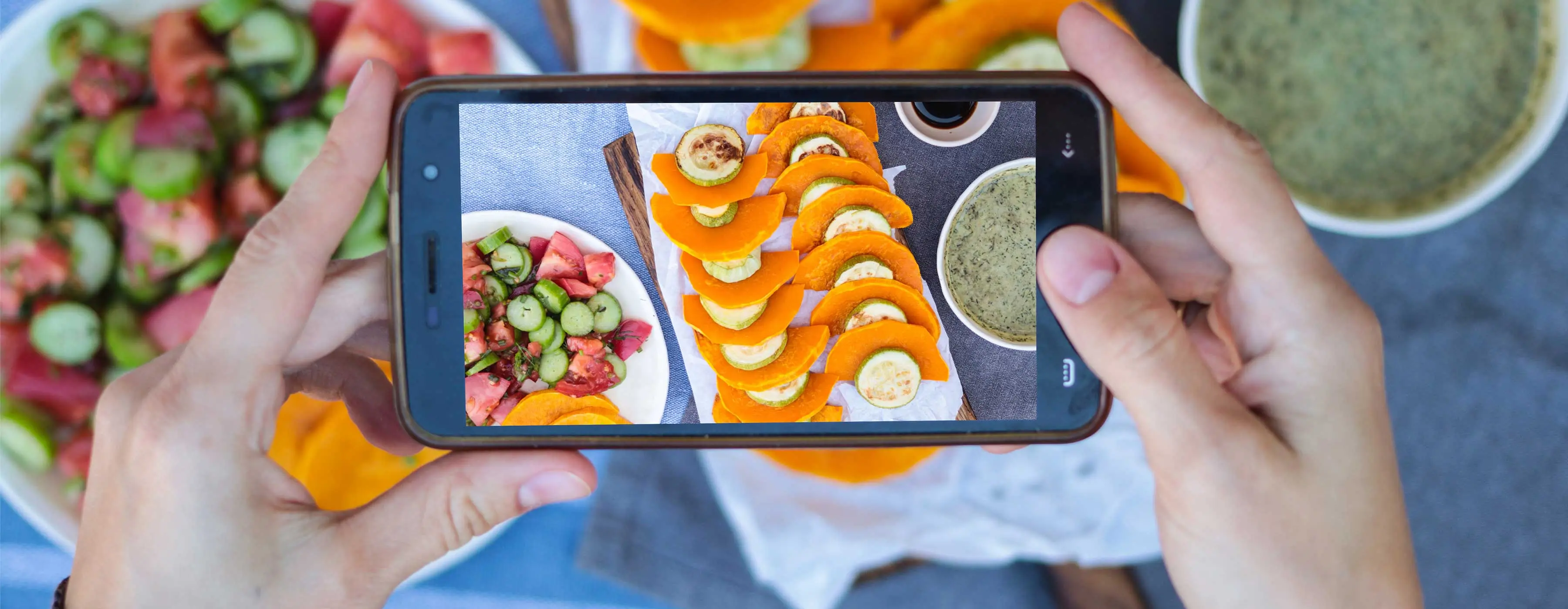 Foto de pessoa fotografando com celular um prato de salada, legumes e patê, com copo de café ao lado.