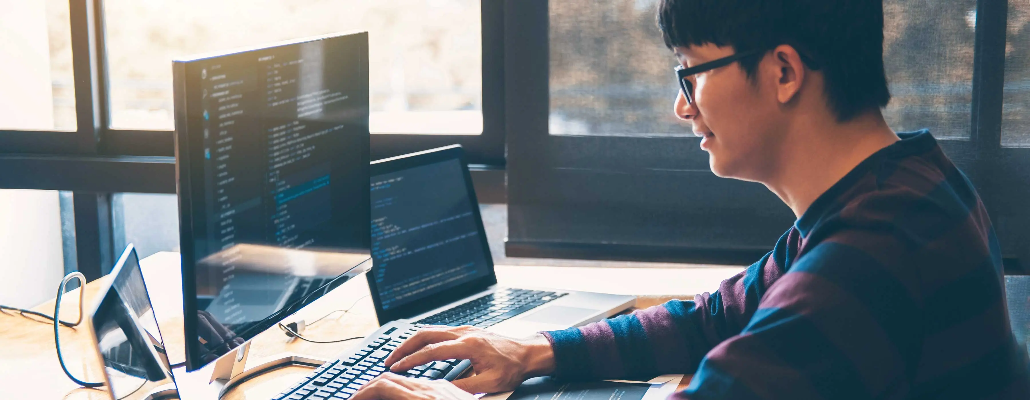 Foto de homem programando em programa JavaScript em computador e dois notebooks em escritório.