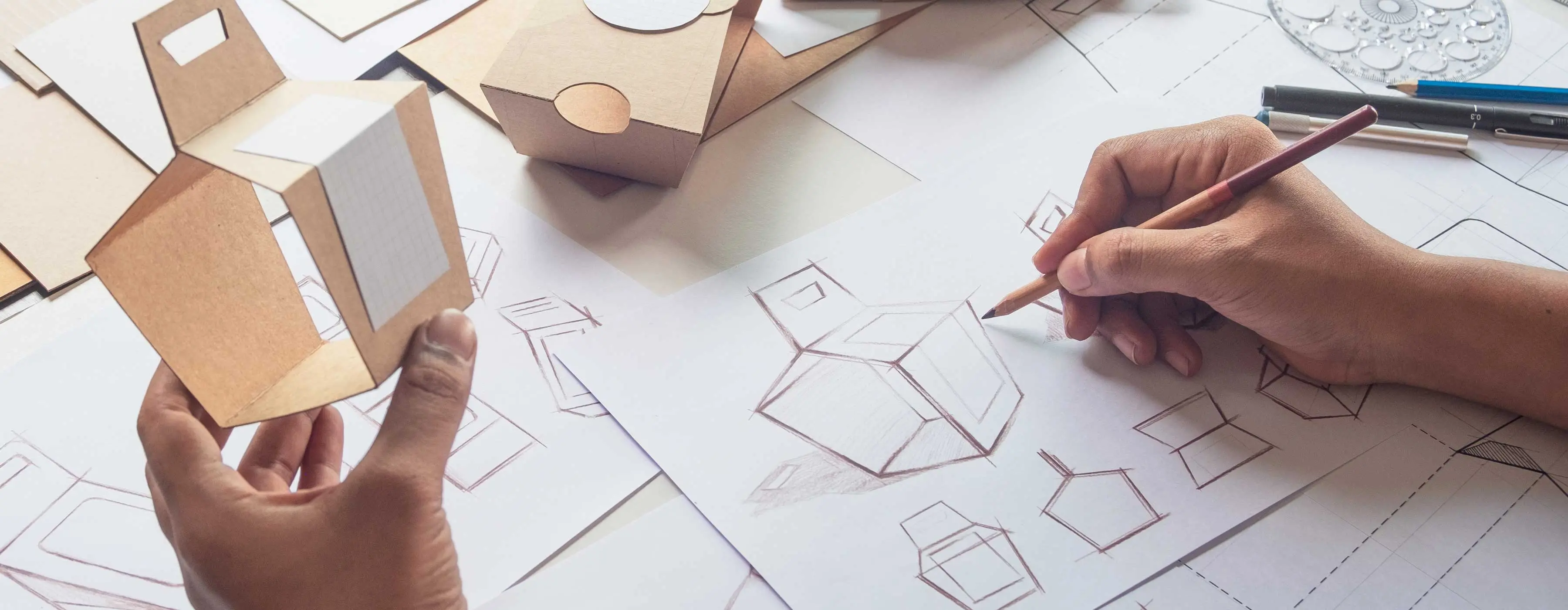 Foto de mão segurando protótipo de caixa de papel enquanto outra mão desenha modelos de caixas em papel.