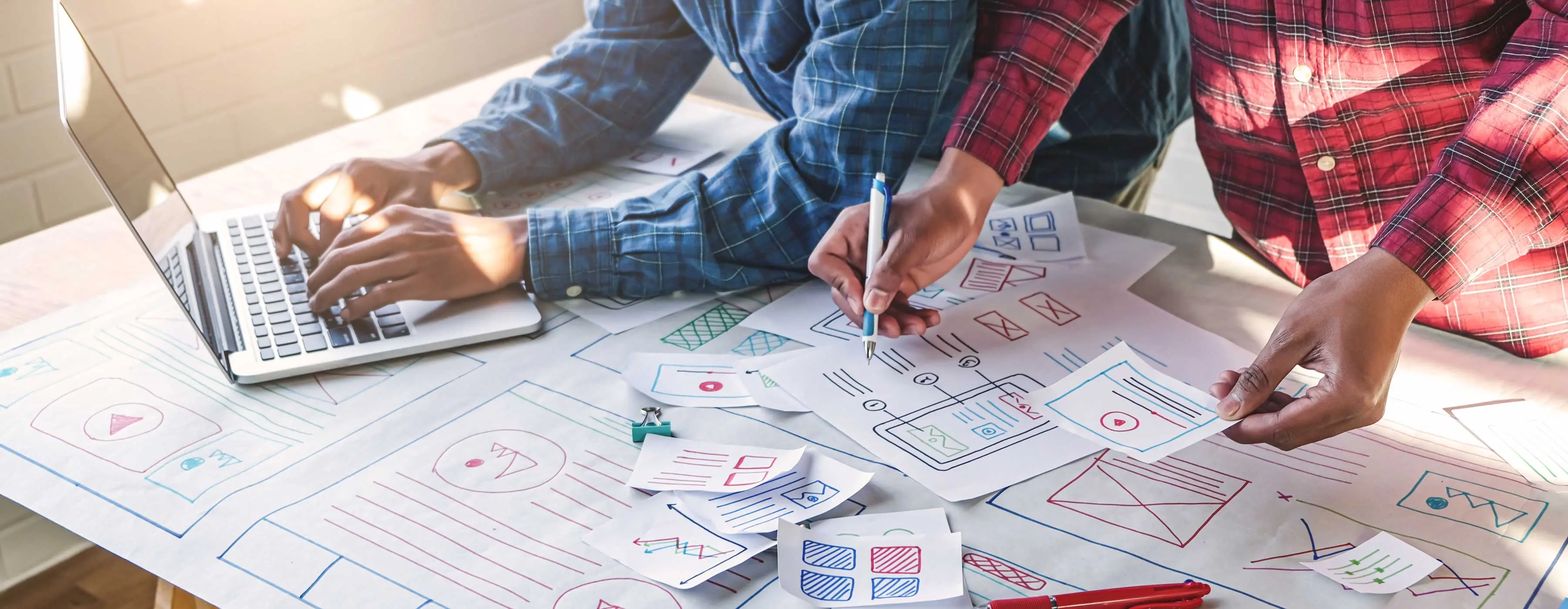 Foto de duas pessoas apoiadas sobre mesa com notebook e papeis com desenhos coloridos de telas e interfaces.
