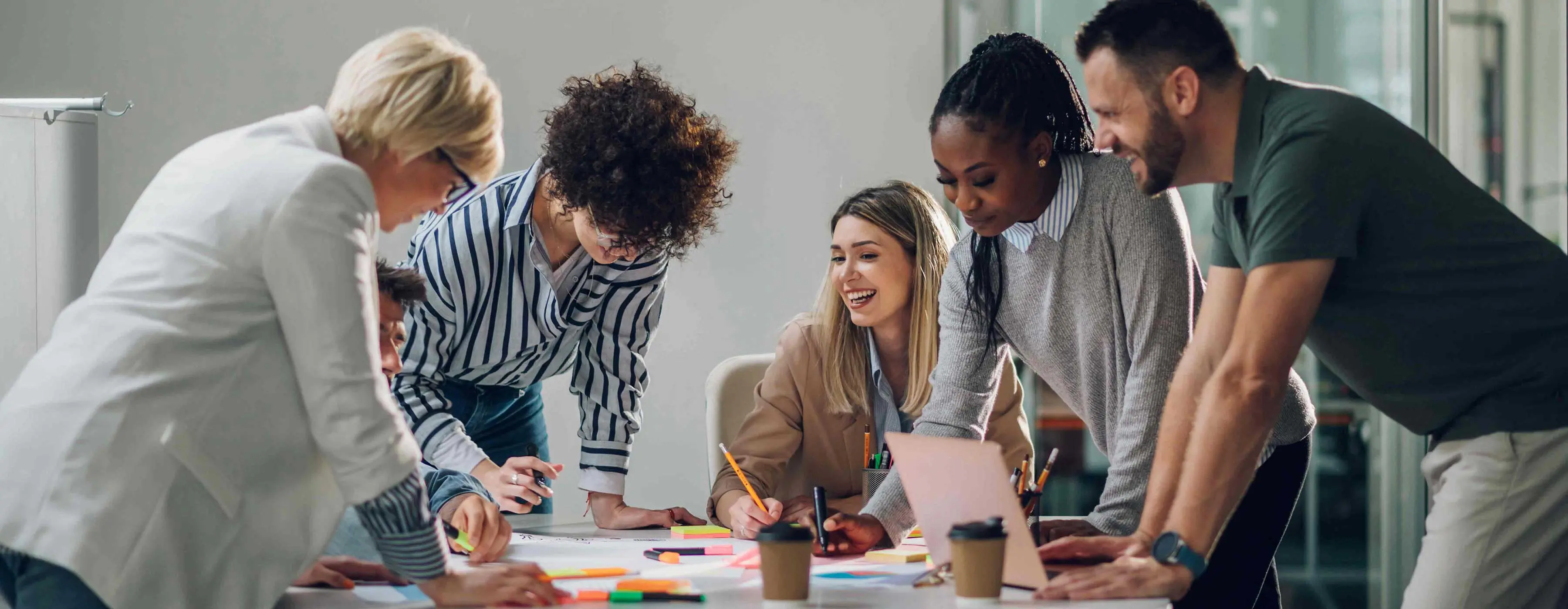 Foto de pessoas reunidas ao redor de mesa com notebook, copos descartáveis, canetas e papéis coloridos.