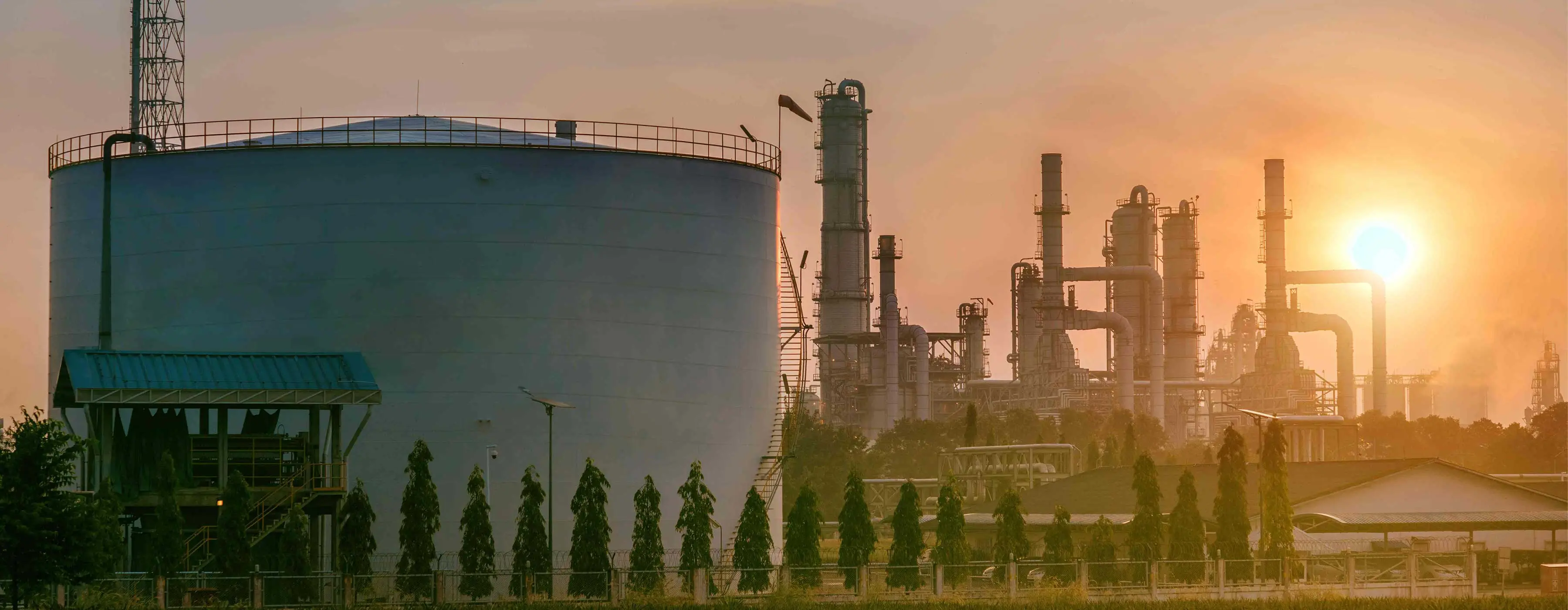 Foto de ambiente externo de refinaria de óleo com árvores ao redor e sol ao fundo.