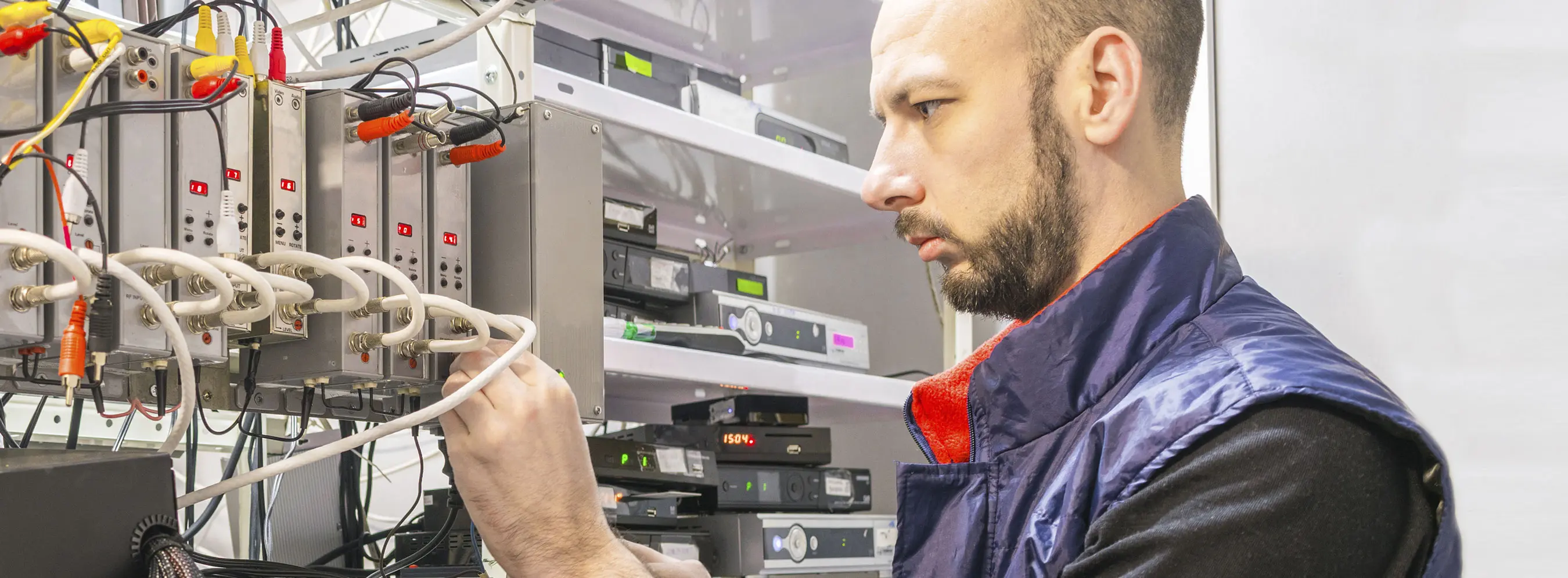 Foto de homem com semblante sério e concentrado, vestido com colete e mexendo em equipamentos de tecnologia.