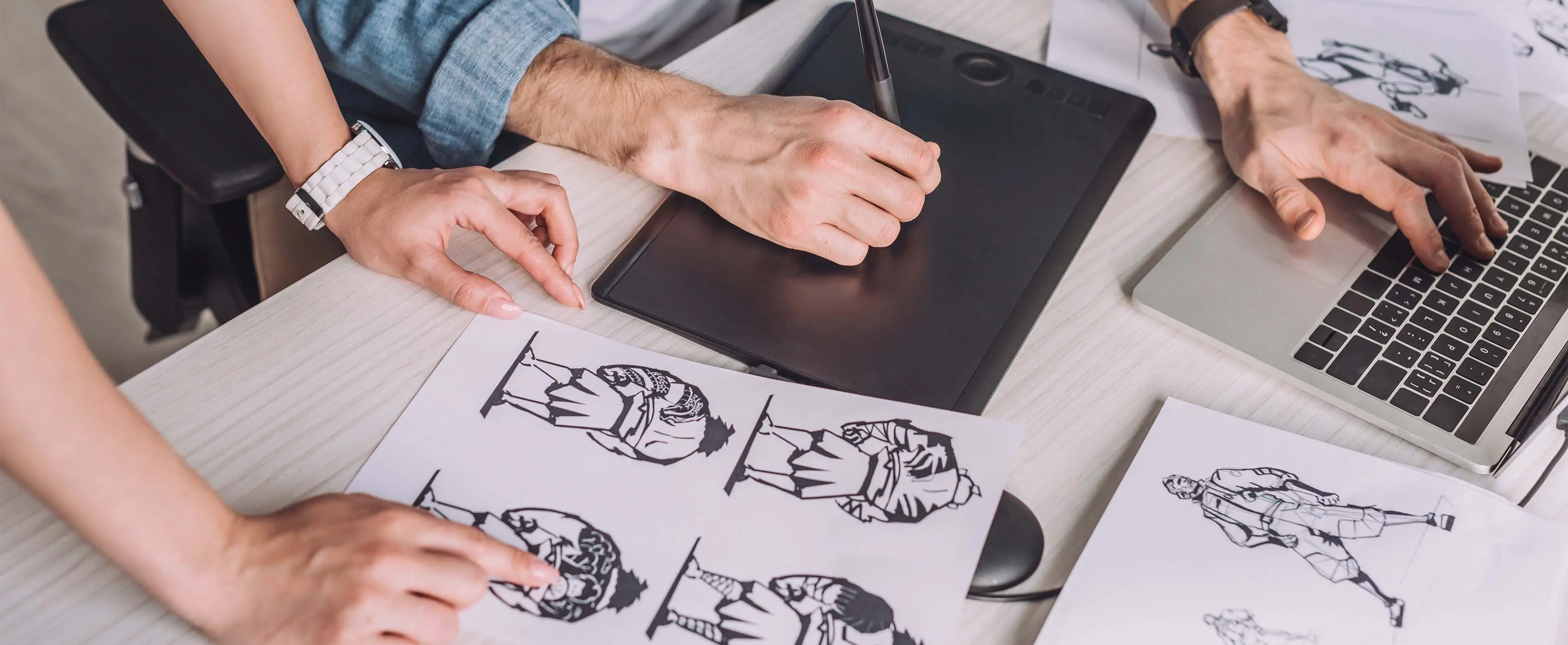 Foto de quatro mãos apoiadas sobre mesa com notebook, mesa e caneta digitalizadoras e ilustrações em papeis.