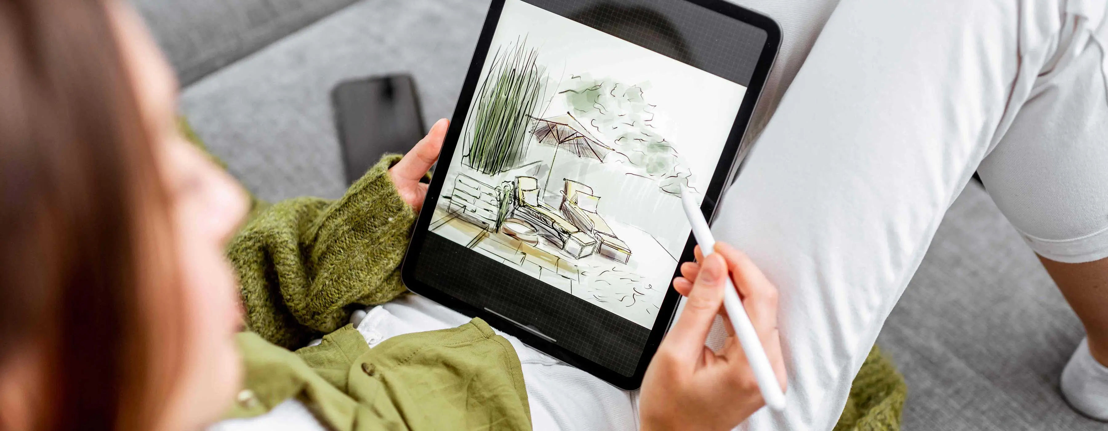foto de uma mulher vestindo blusa na cor verde, segurando um tablet no colo e desenhando um ambiente com cadeiras de descanso, guarda-sol e plantas.