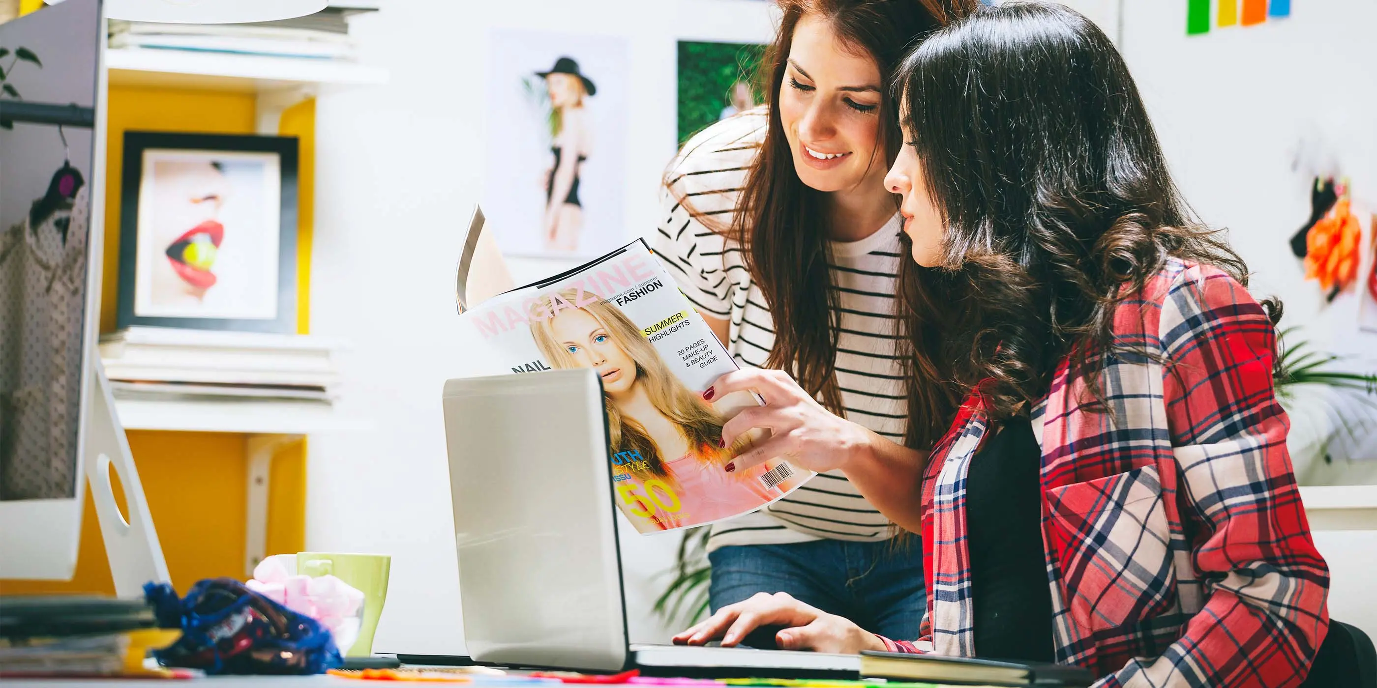 Foto de duas mulheres olhando para revista de moda em frente a notebook e com imagens e referências de moda ao redor.