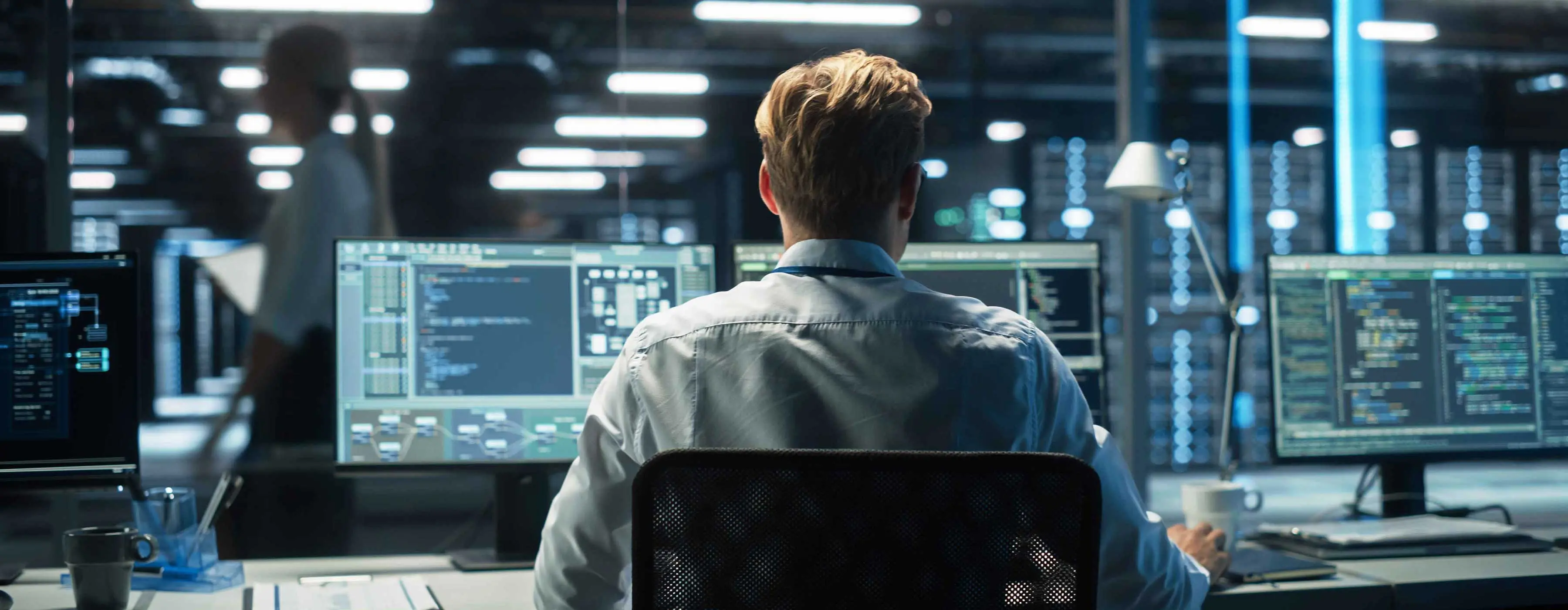 Foto de um homem loiro vestindo camisa branca, de costas, sentado em frente a monitores exibindo programa de dados, em um espaço repleto de equipamentos e outras telas de computador.