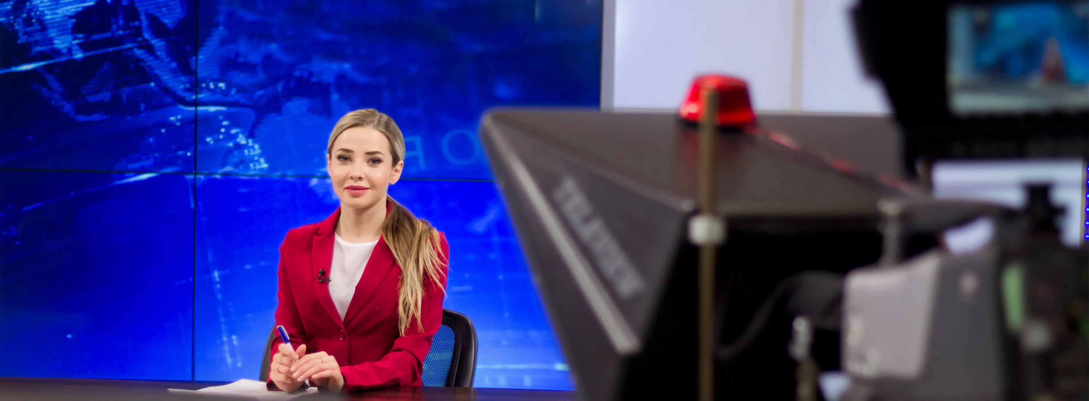 Foto de mulher sentada atrás de uma bancada de telejornal em estúdio de TV olhando sorridente para câmera de vídeo.